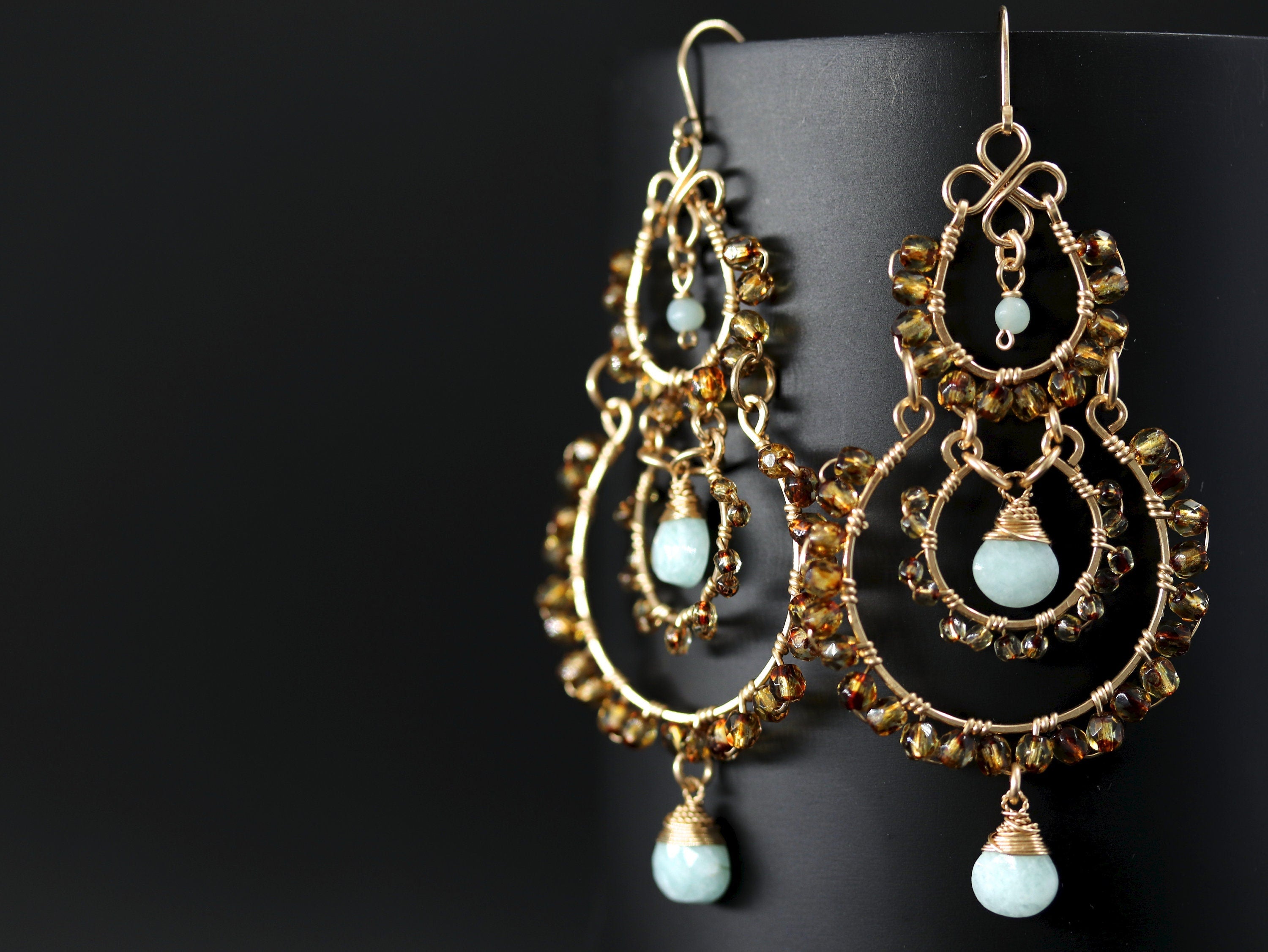 May include: Gold-toned chandelier earrings with multiple tiers. Each tier is adorned with amber-colored beads and features a teardrop-shaped, pale blue gemstone pendant. The earrings have a delicate, wire-wrapped design, creating an elegant and intricate look.