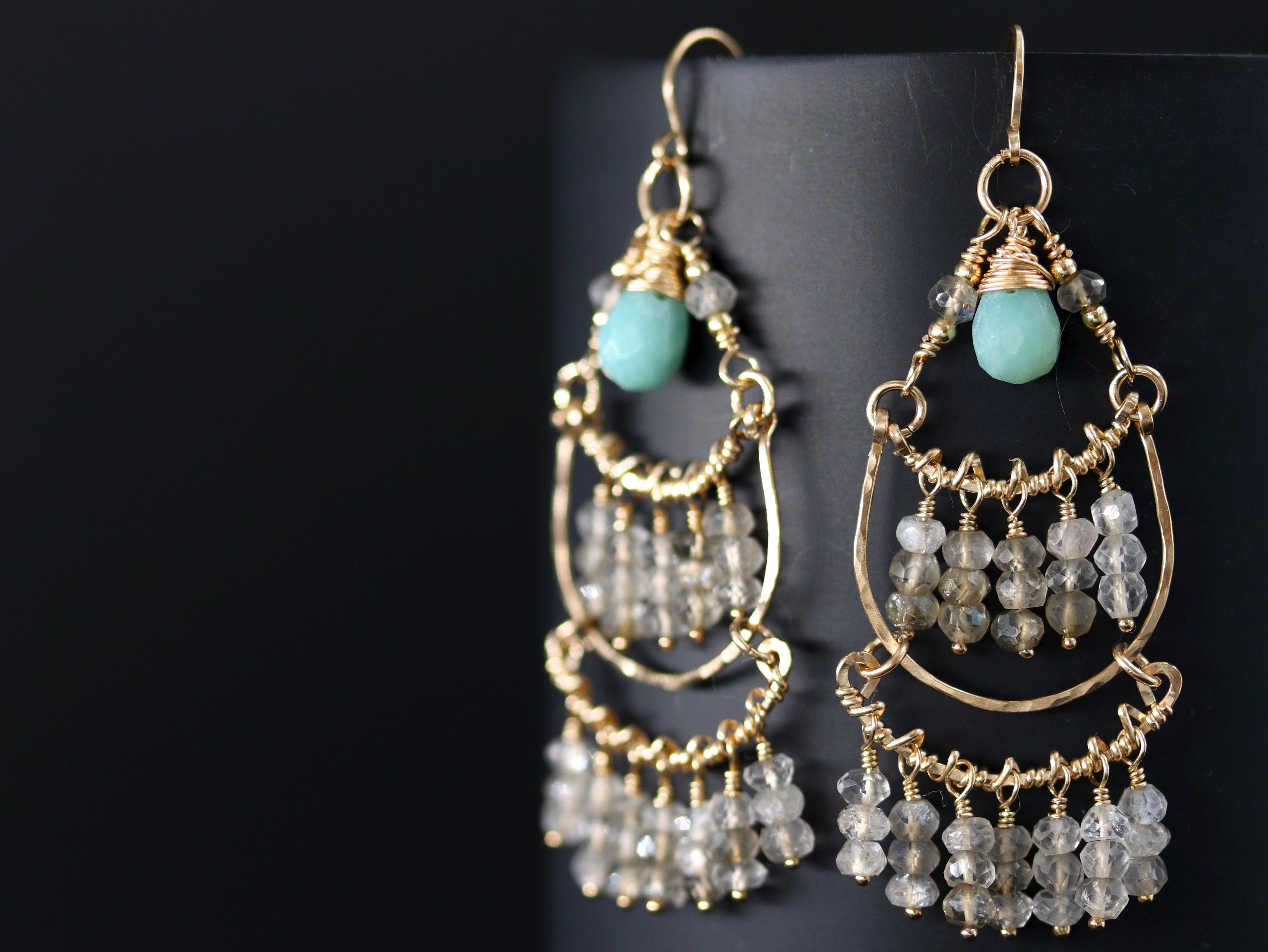 May include: Gold dangle earrings with a turquoise gemstone and clear faceted beads. The earrings have a layered design with multiple tiers of beads.