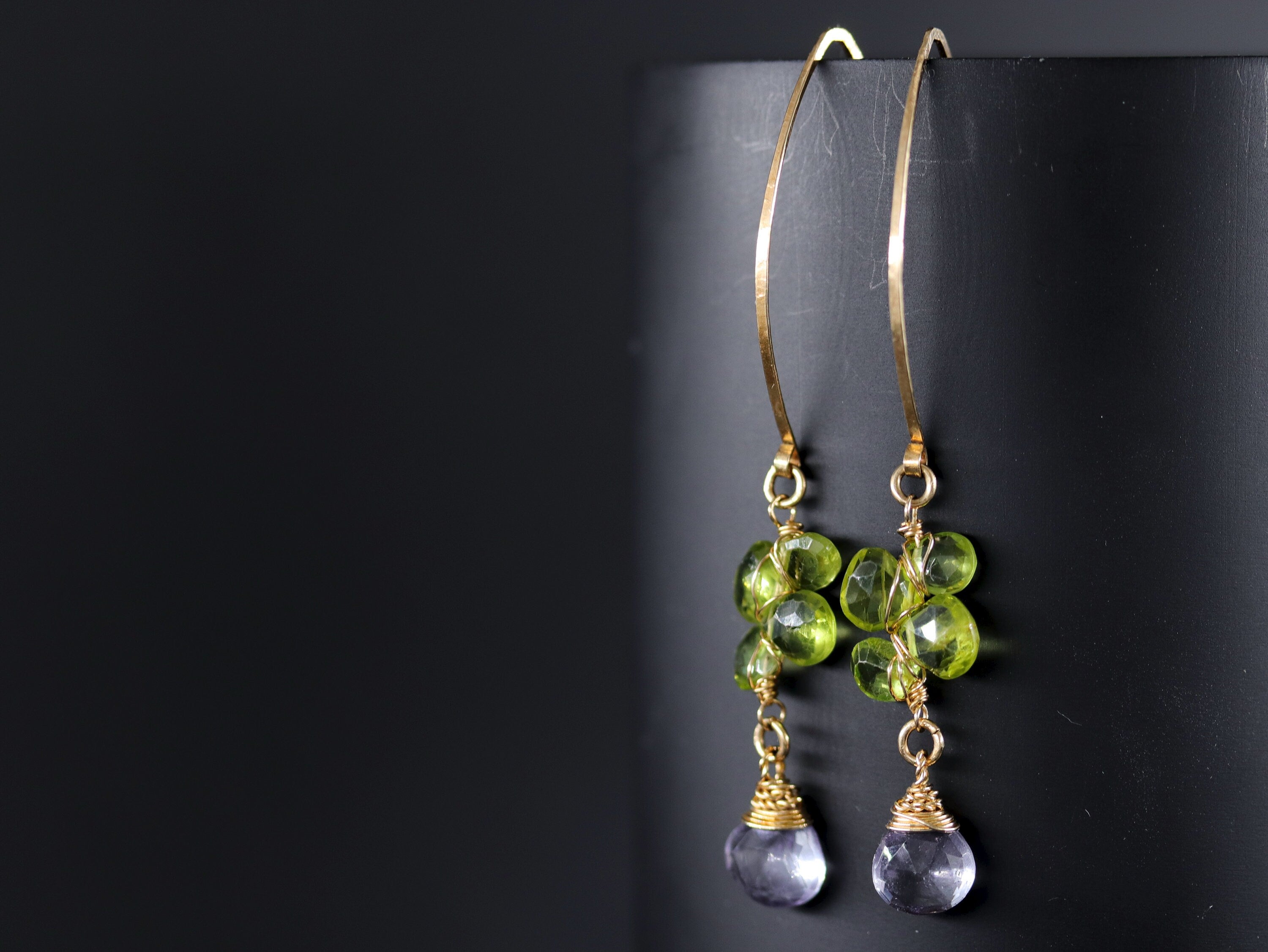 May include: Gold earrings with green and purple gemstones. The earrings feature a cluster of green gemstones and a single purple gemstone dangling from the bottom.