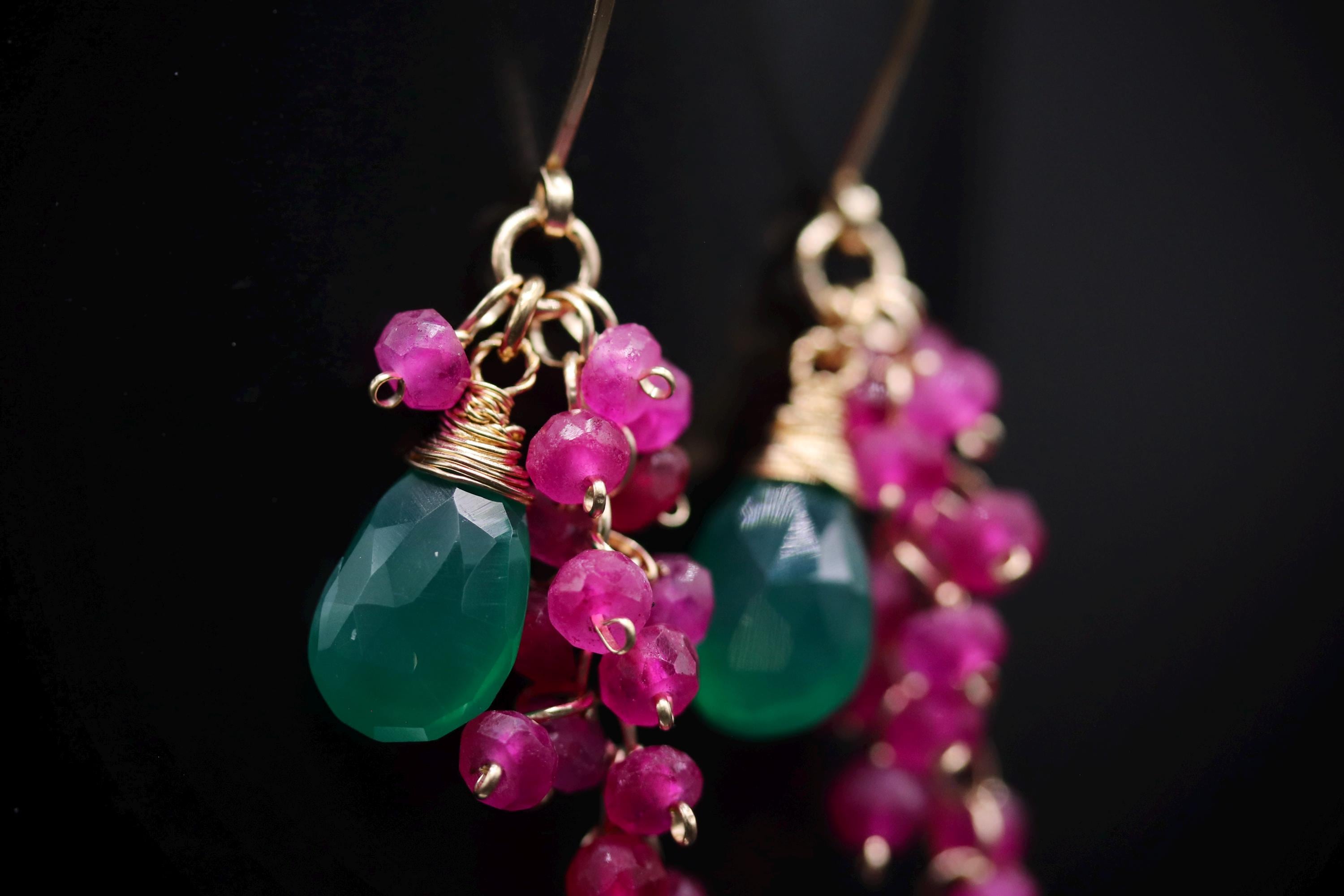 A close-up view of a pair of earrings with green and pink gemstones and dangling charms.