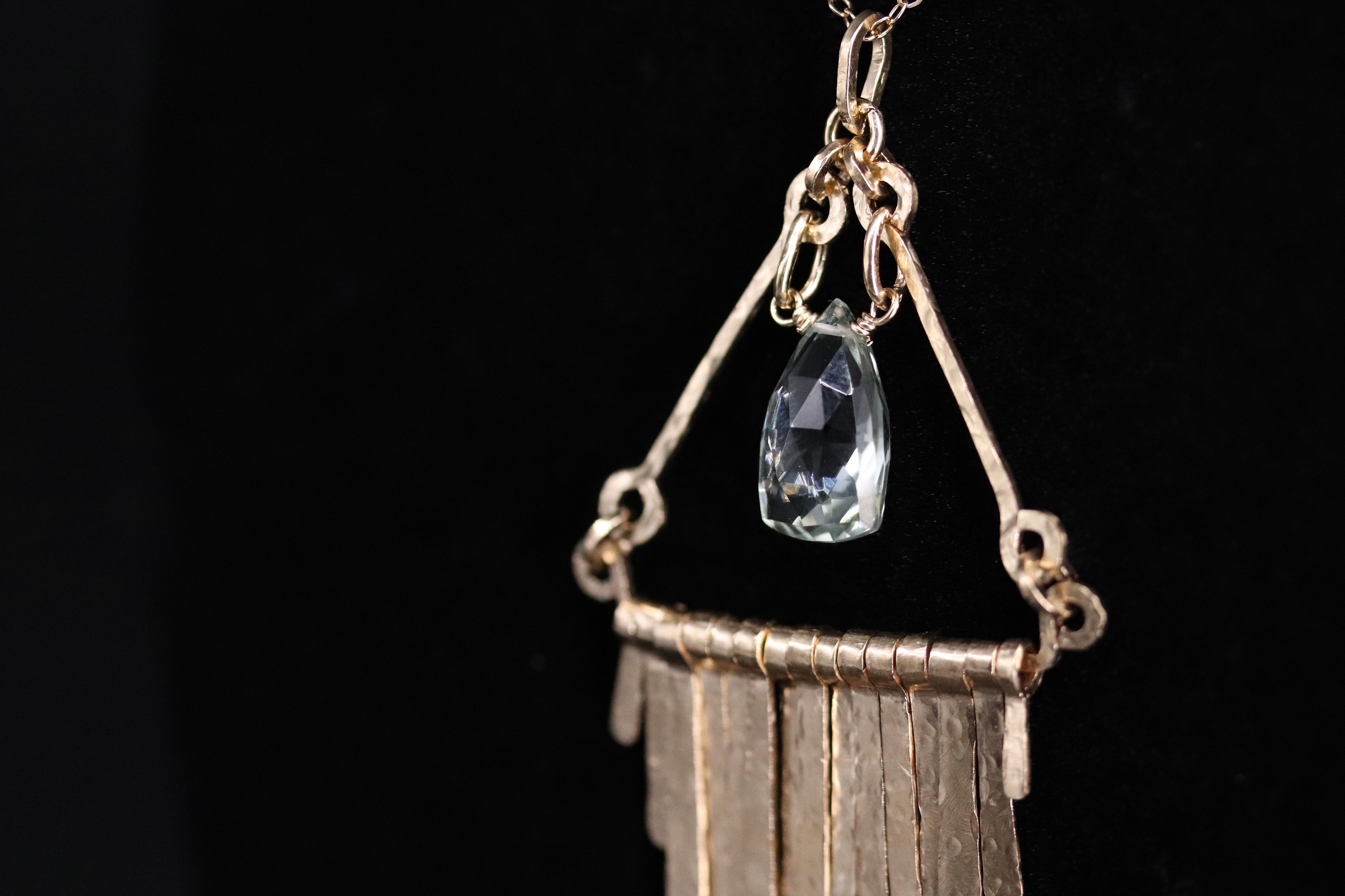 A close-up view of a delicate necklace with a clear gemstone pendant, hanging against a black background.