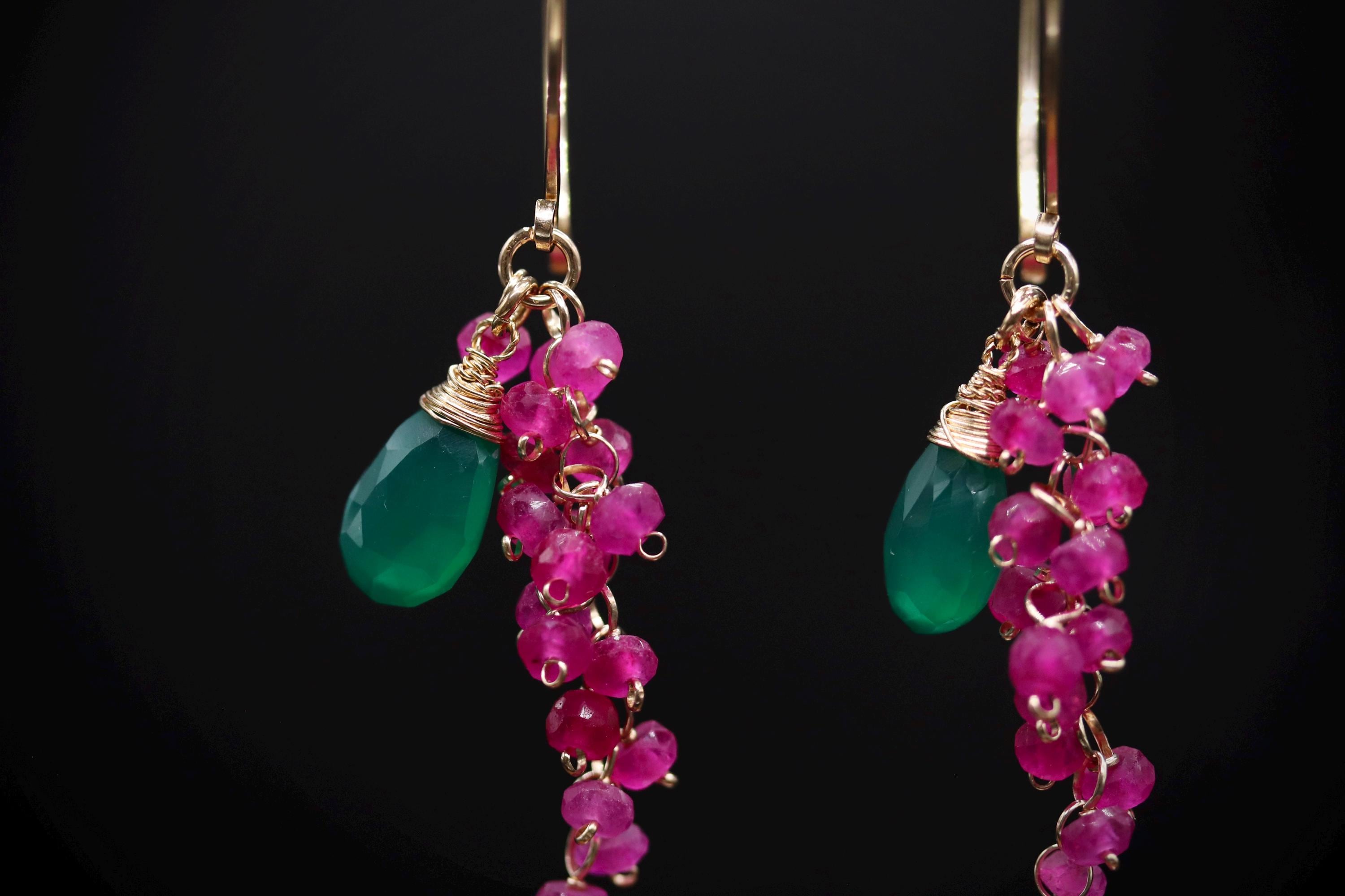 A pair of earrings with green gemstones and pink gemstone charms hanging from them against a black background.