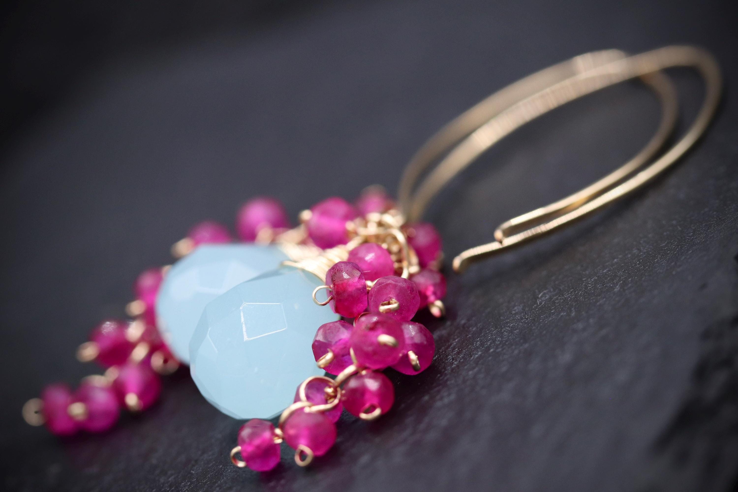 A close-up image of a pair of earrings with a combination of blue and pink gemstones, as well as gold-colored metal earring hooks.