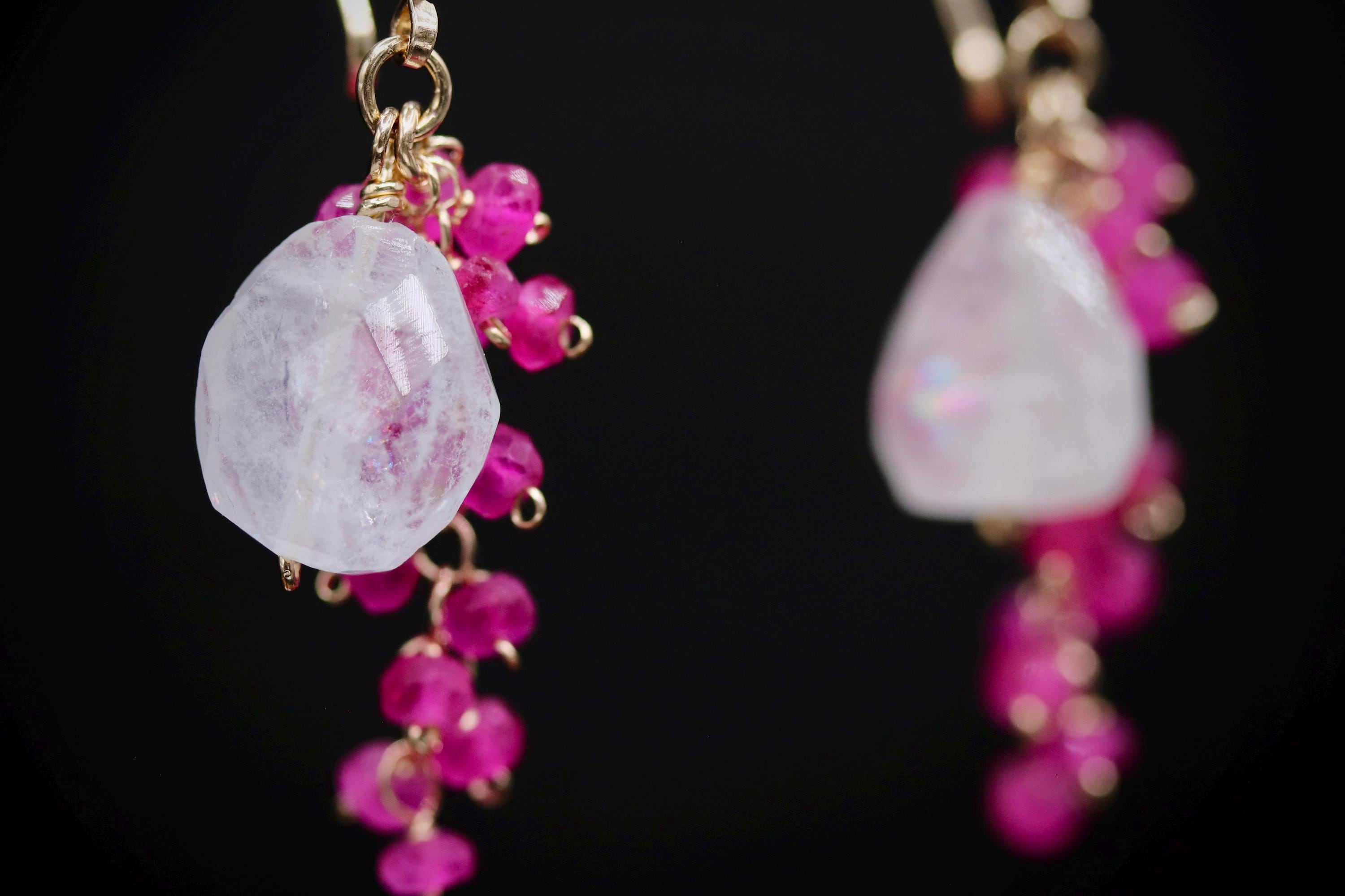 A close-up view of a pair of earrings with a clear gemstone and a cluster of small, round, pink beads hanging from the earrings.