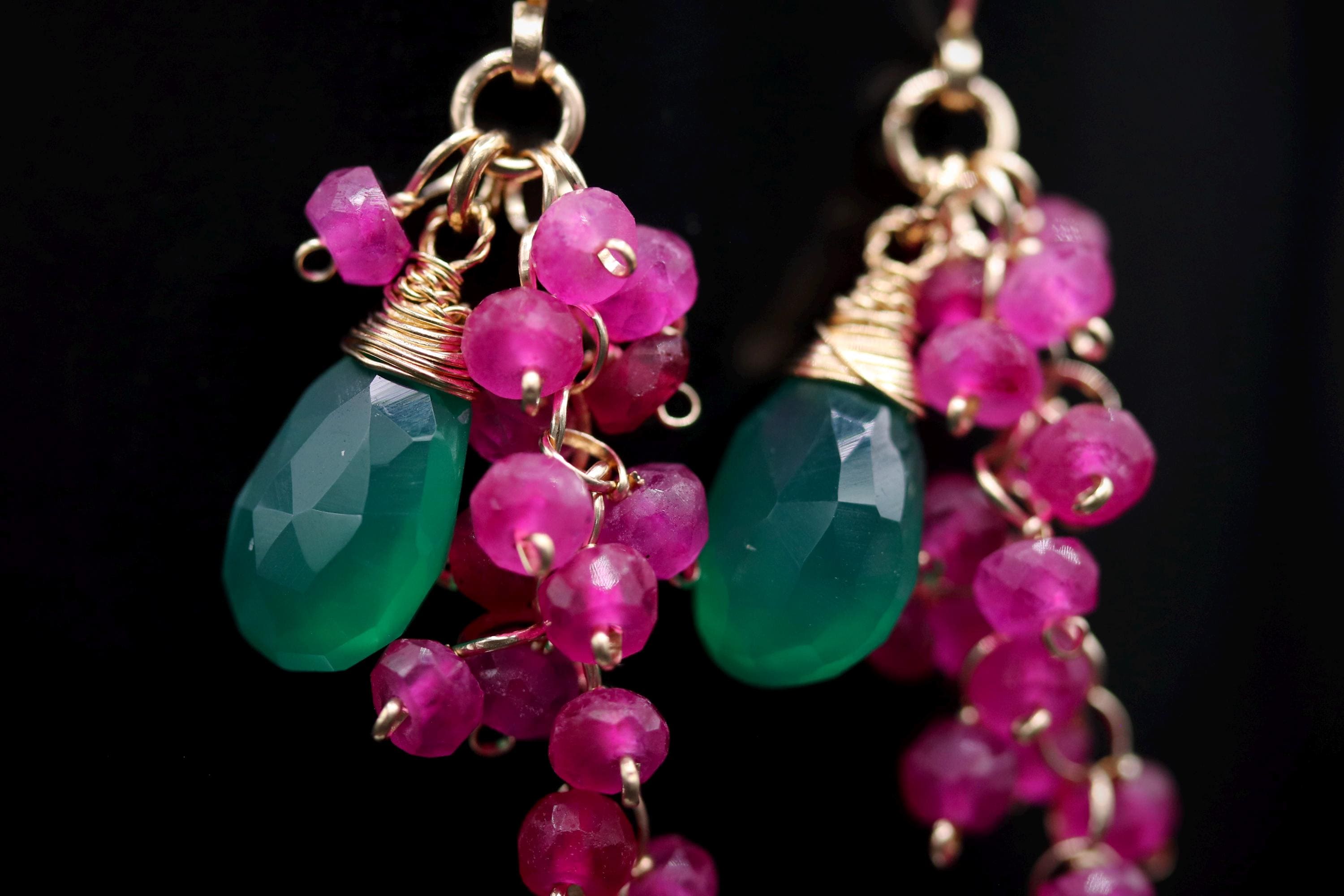 A close-up view of a pair of earrings with green and pink gemstones and dangling beads.