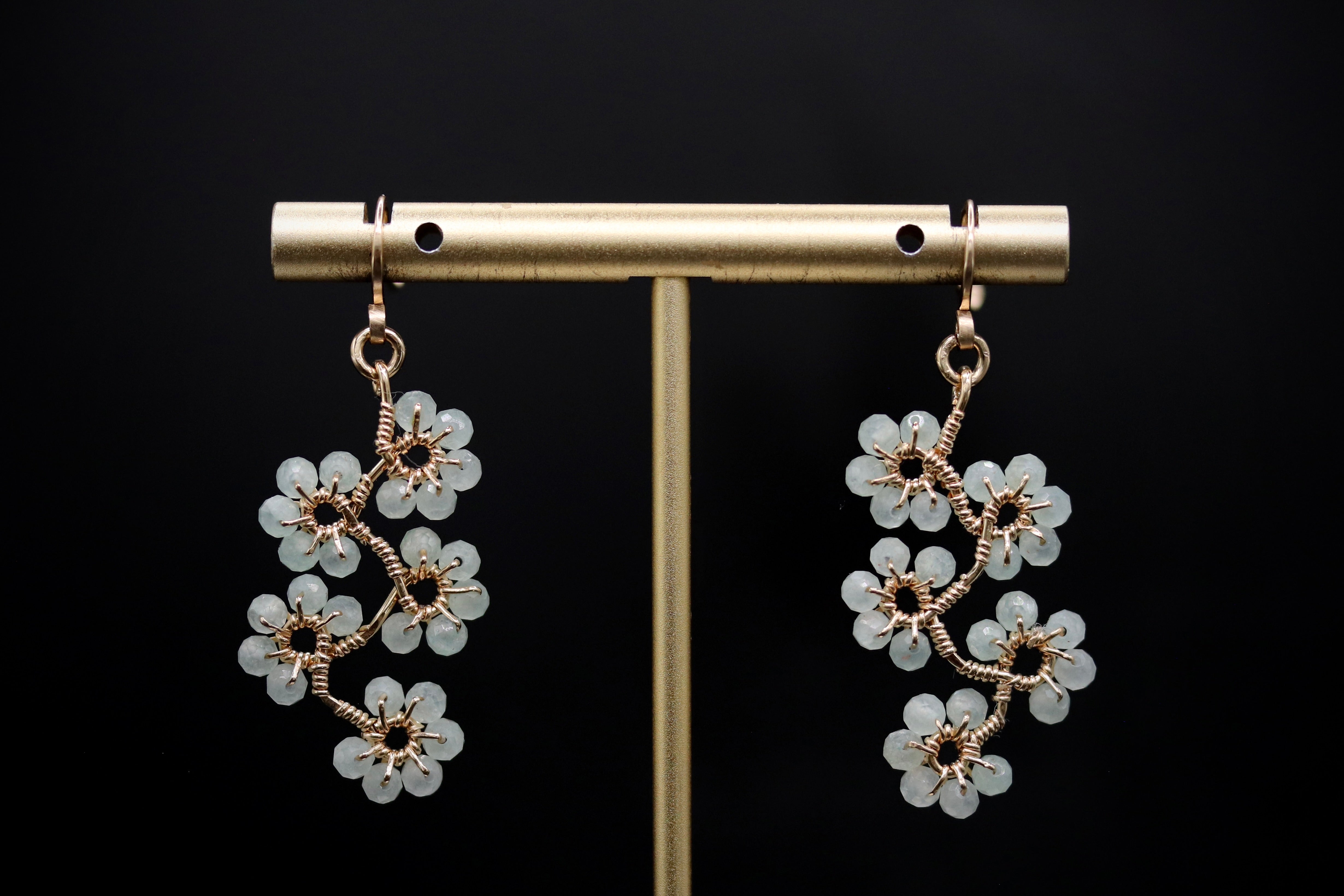 A pair of elegant earrings with white flower-like designs hanging from a gold metal stand against a black background.