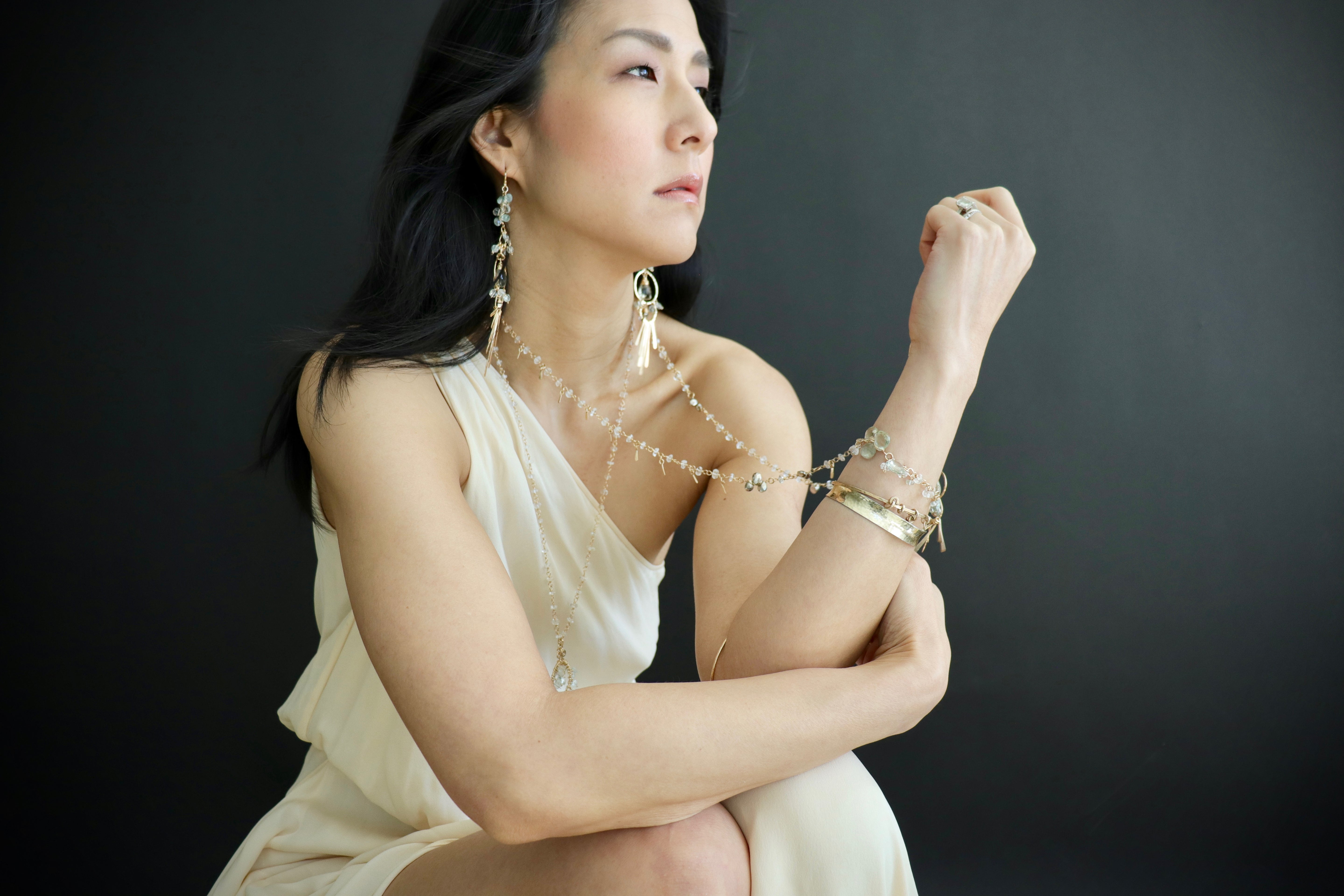 A woman in a white dress poses with her arm raised, wearing multiple pieces of jewelry, including earrings and bracelets.