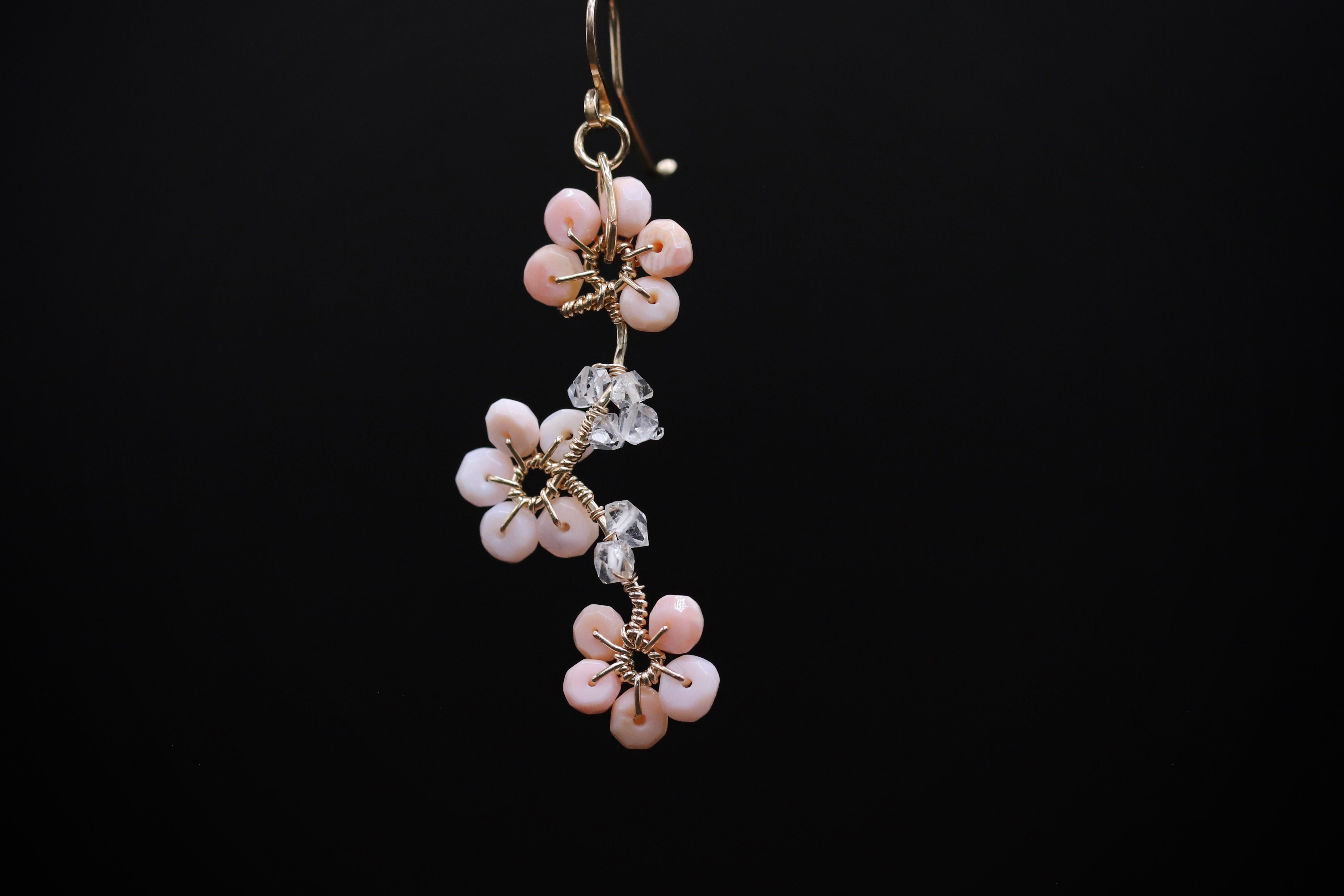 A delicate earring with a floral design, featuring small pink flowers and clear gemstones, is suspended against a black background.