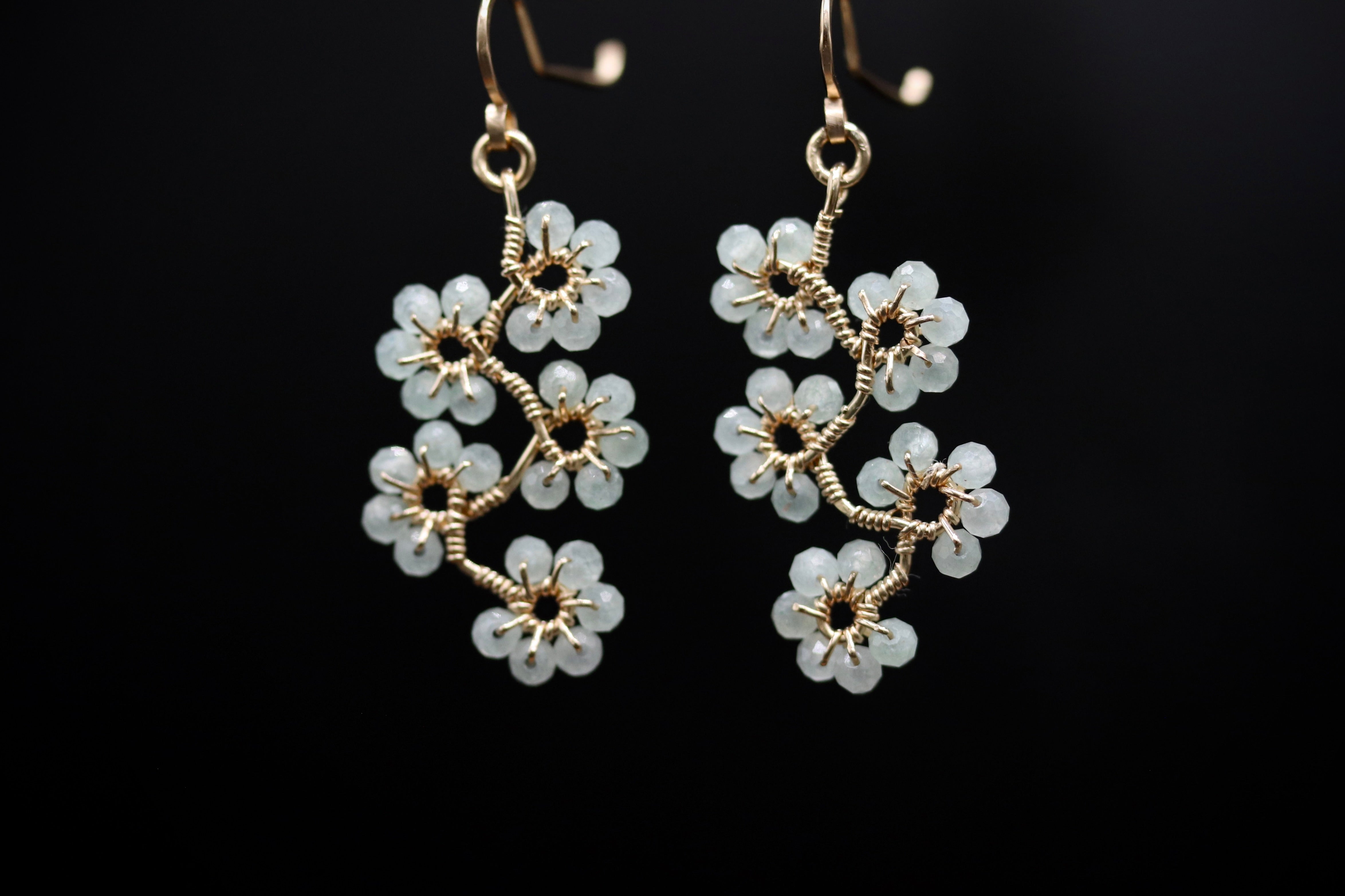 A pair of elegant gold earrings with white flower-shaped charms hanging from the hooks against a black background.