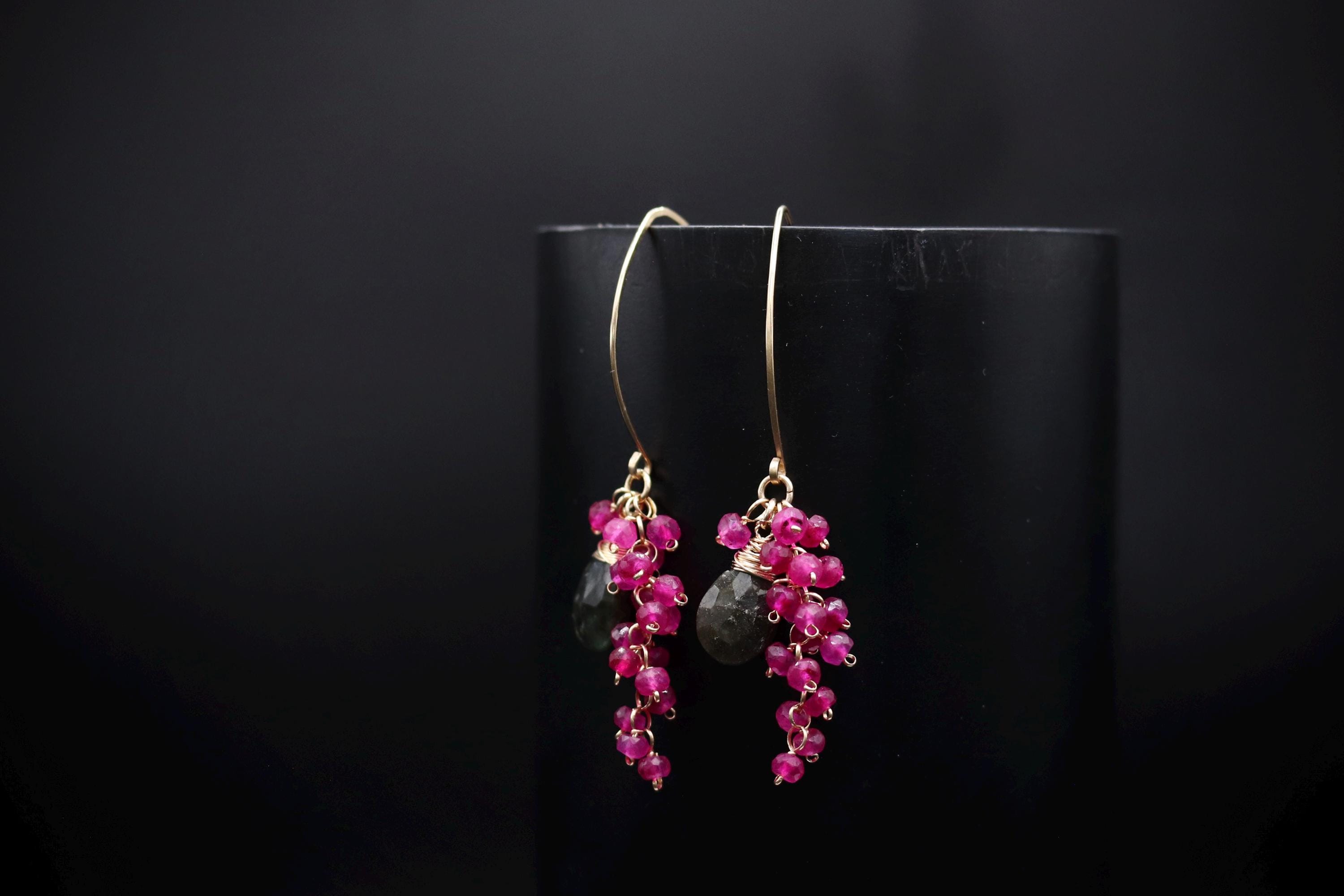A pair of earrings with a black background and a gold hook, featuring a cluster of pink gemstones.