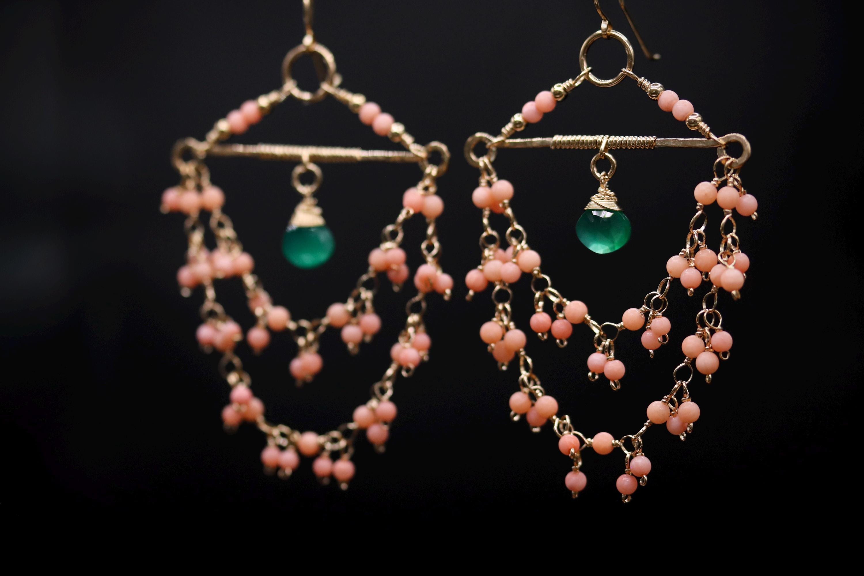 A pair of earrings with a green gemstone and pink beads hanging from a decorative frame against a dark background.