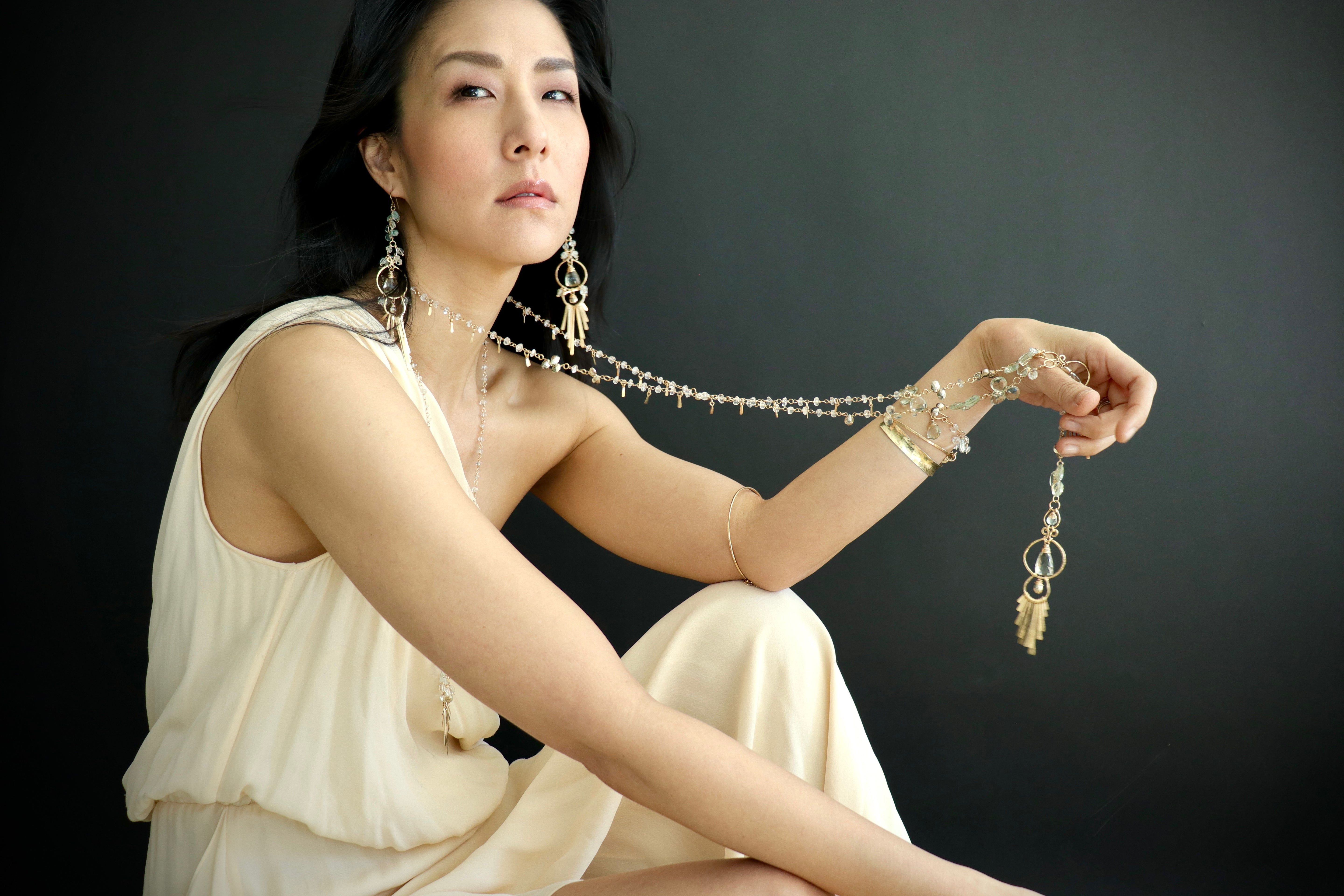 A woman in a white dress poses with a long necklace and earrings against a dark background.