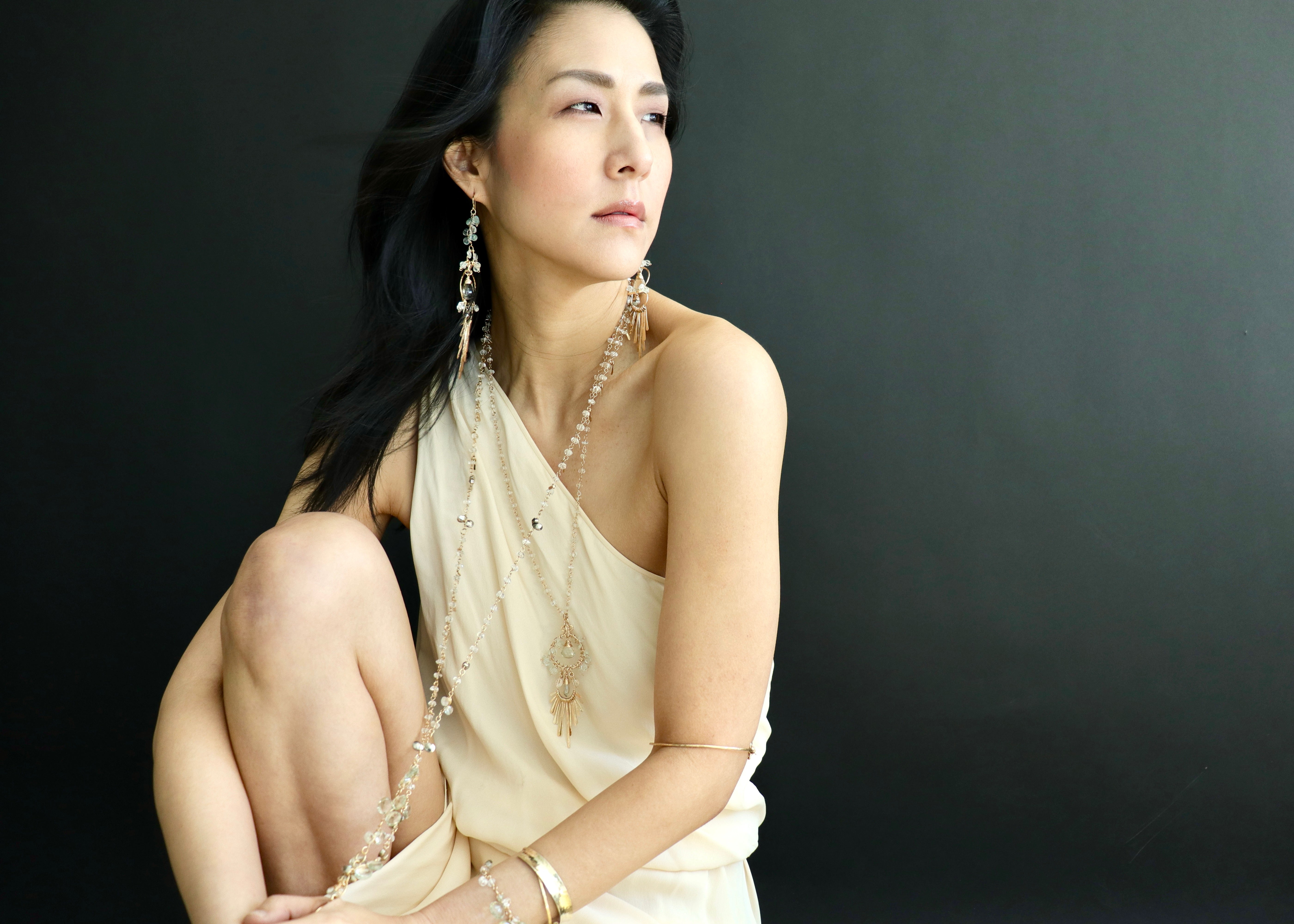A woman in a white dress and earrings is posing against a dark background, looking off to the side with a thoughtful expression.