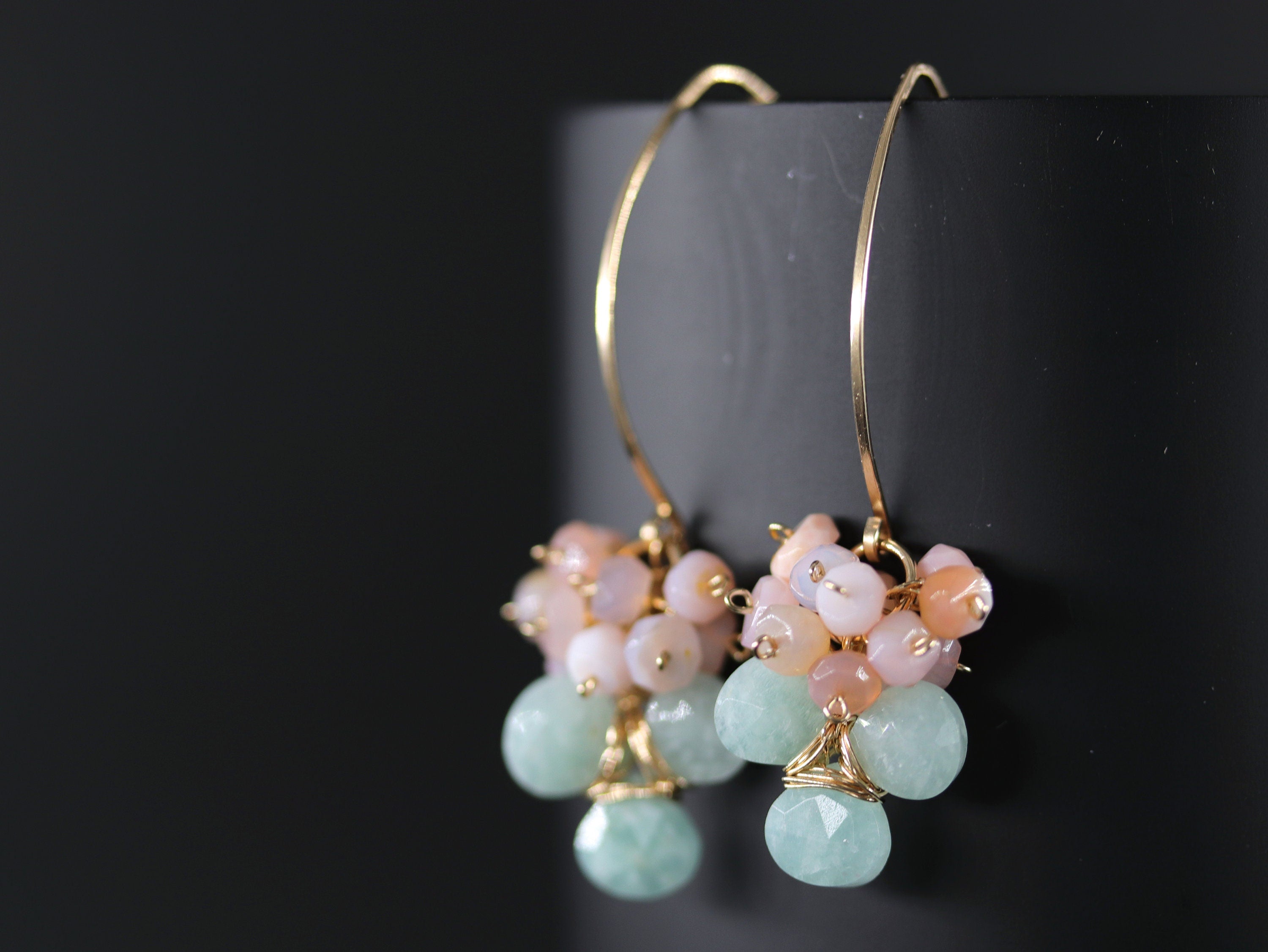 May include: Gold hoop earrings with a cluster of light pink and peach gemstones and a cluster of light green gemstones hanging from each hoop.