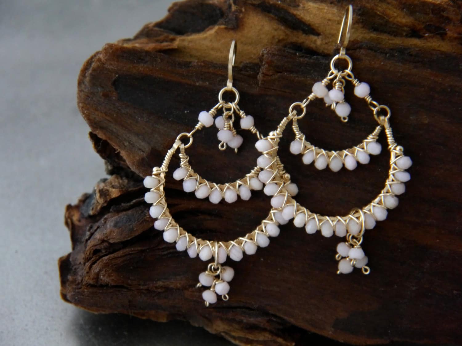 May include: Gold-toned chandelier earrings with a delicate design. The earrings feature a crescent shape adorned with small, pale pink beads. The beads are strung along the wire frame, creating a layered effect. The earrings have a hook closure.