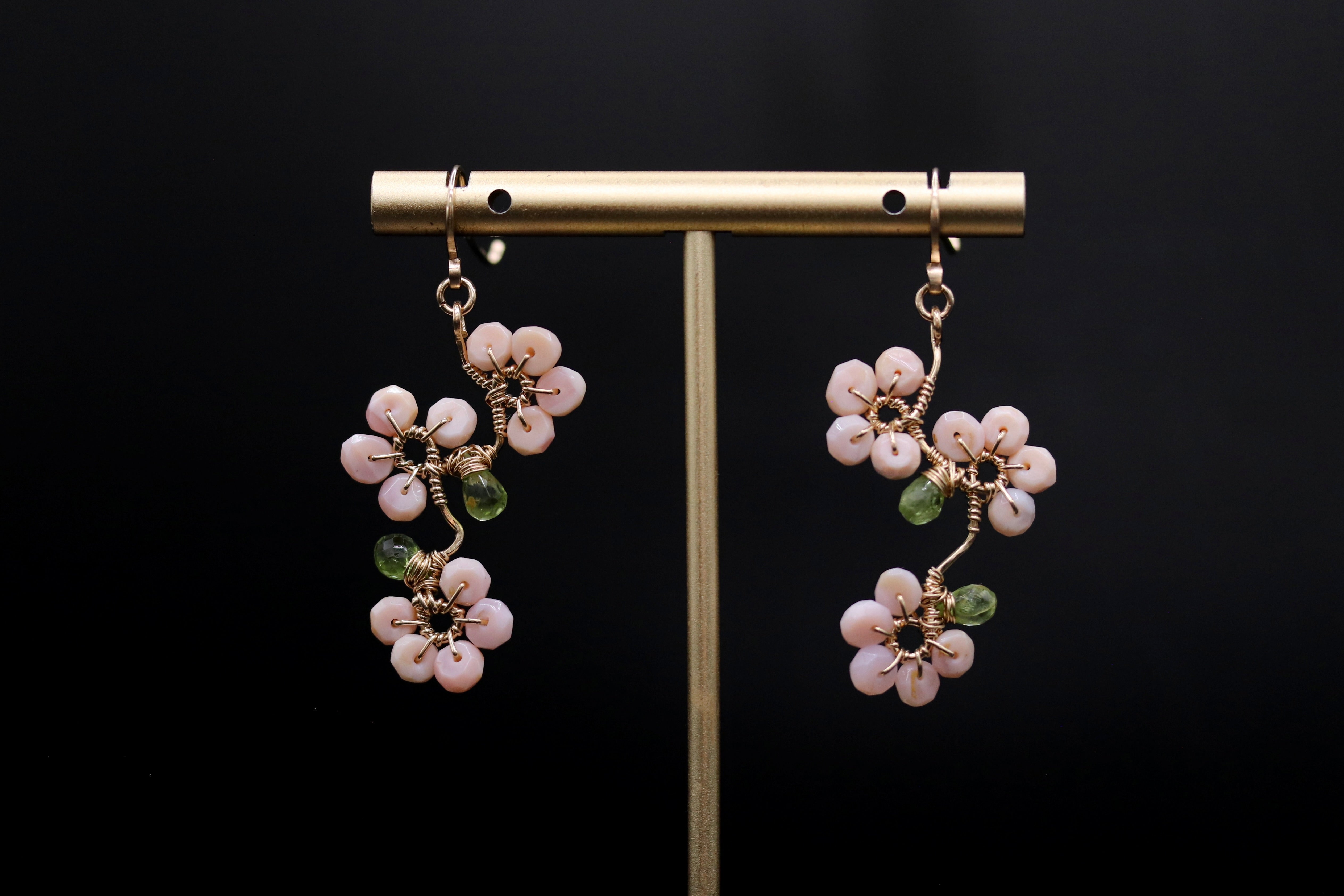 A pair of delicate earrings with pink and green floral designs, displayed on a gold stand against a black background.