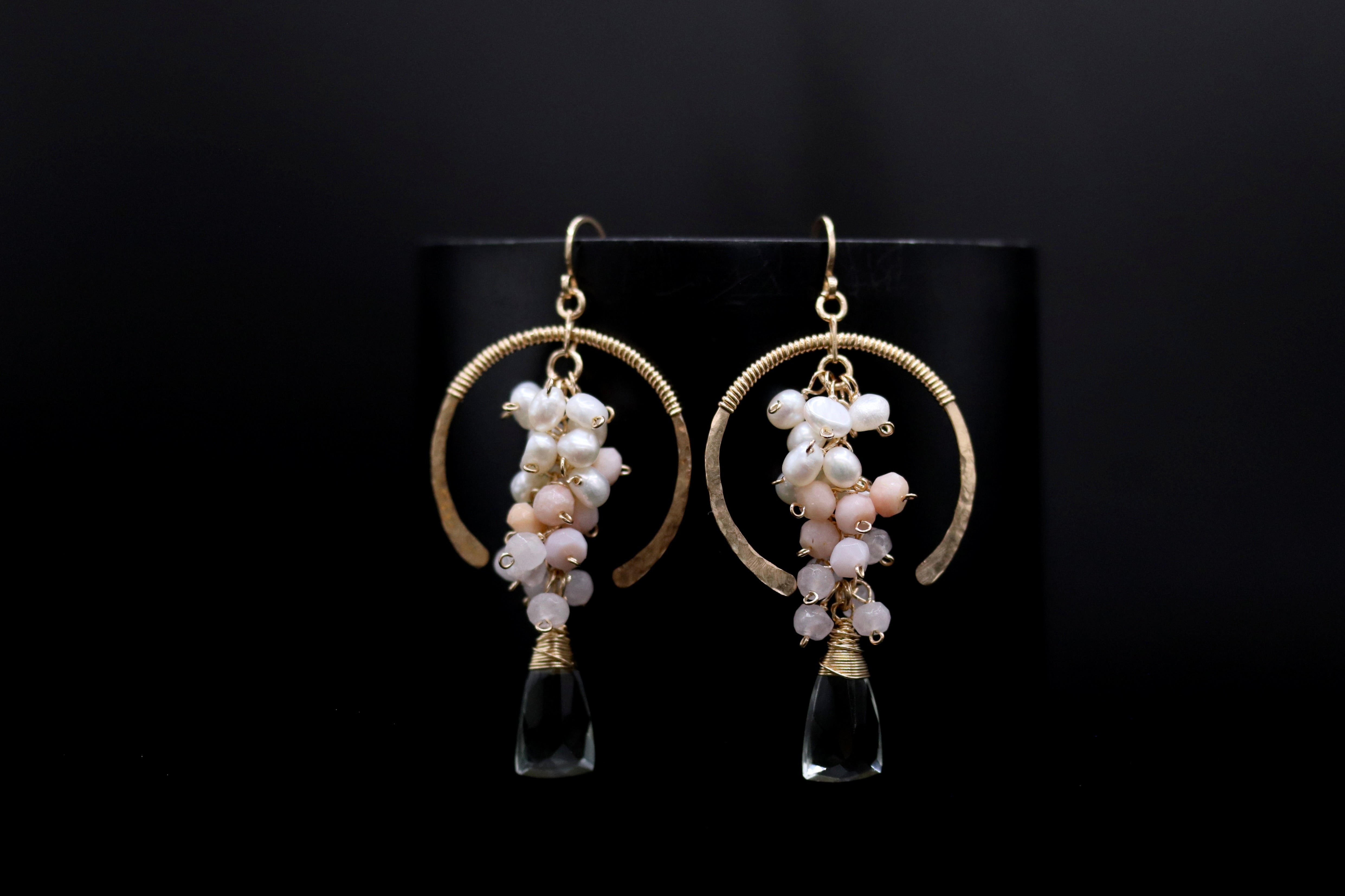 A pair of elegant earrings with a black background. The earrings have a gold hoop design and are adorned with white and pink pearls, as well as clear gemstone accents.