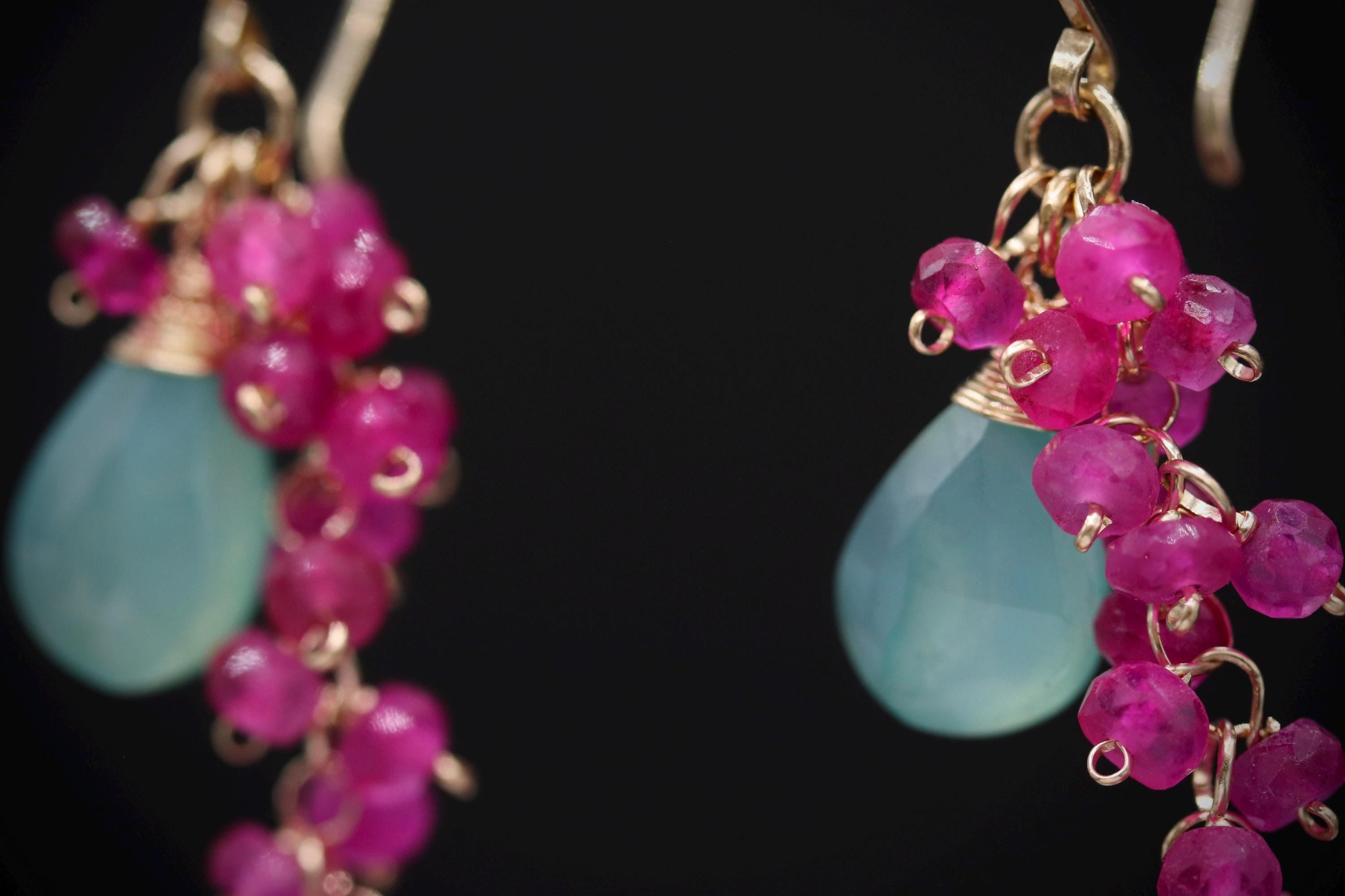 A close-up view of a pair of earrings with a combination of green and pink gemstones, as well as small, dangling beads.