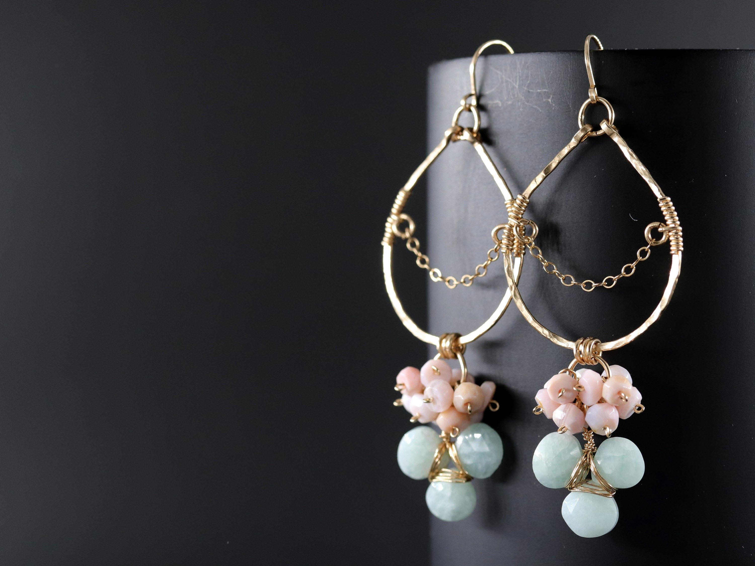May include: Gold-toned dangle earrings with a teardrop design. The earrings feature clusters of pink and light blue beads, connected by delicate gold chains and wire. The background is a dark, solid color.