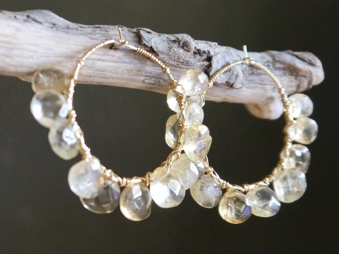May include: A pair of gold hoop earrings with dangling, faceted, yellow-toned gemstones. The earrings are displayed on a piece of driftwood against a dark background. The gemstones are teardrop-shaped and vary in size.
