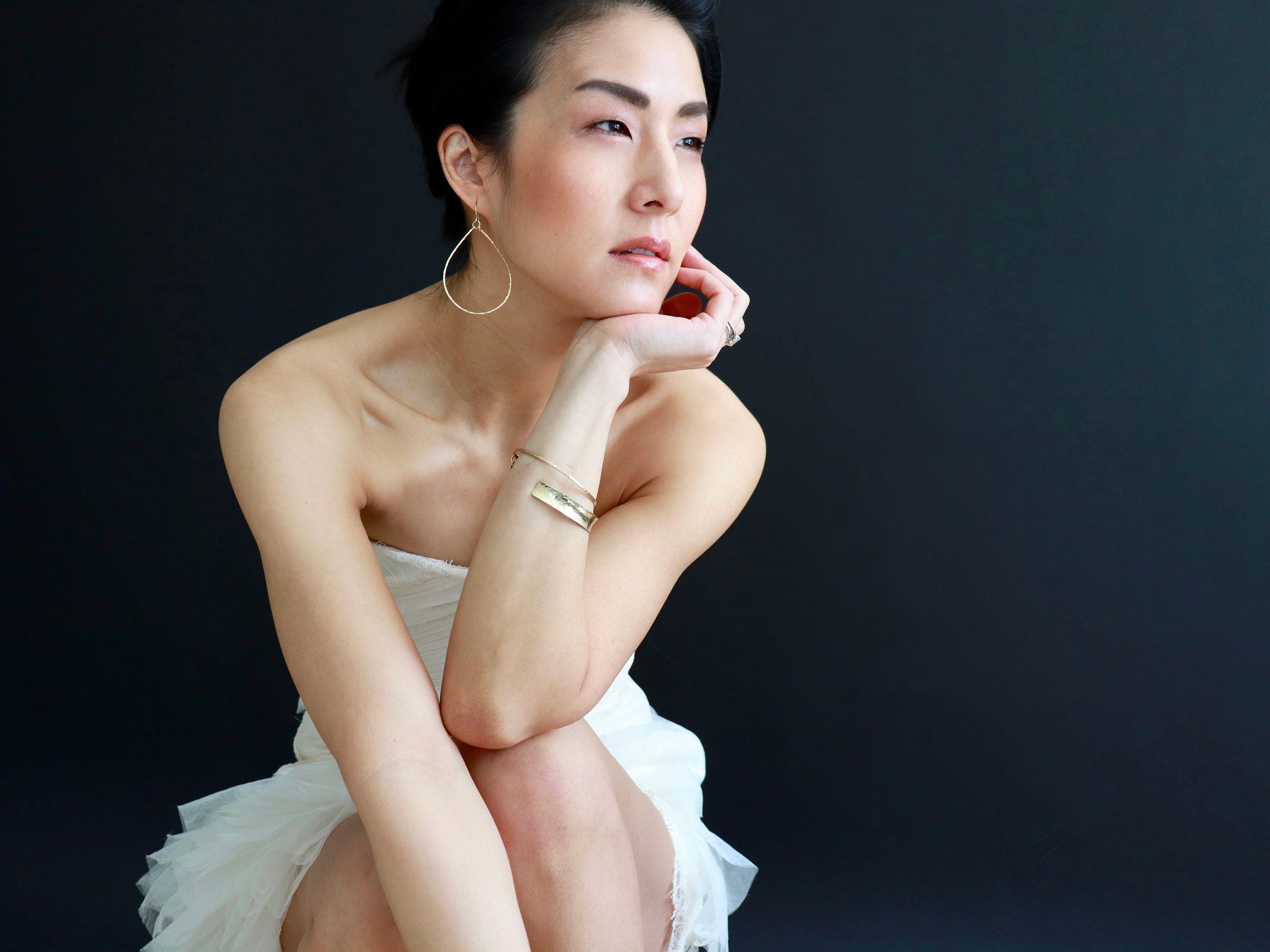 May include: A woman wearing a white strapless dress with a delicate lace trim, sitting with her legs crossed. She is wearing gold earrings and a gold bracelet. Her hair is pulled back and she is looking off to the side.