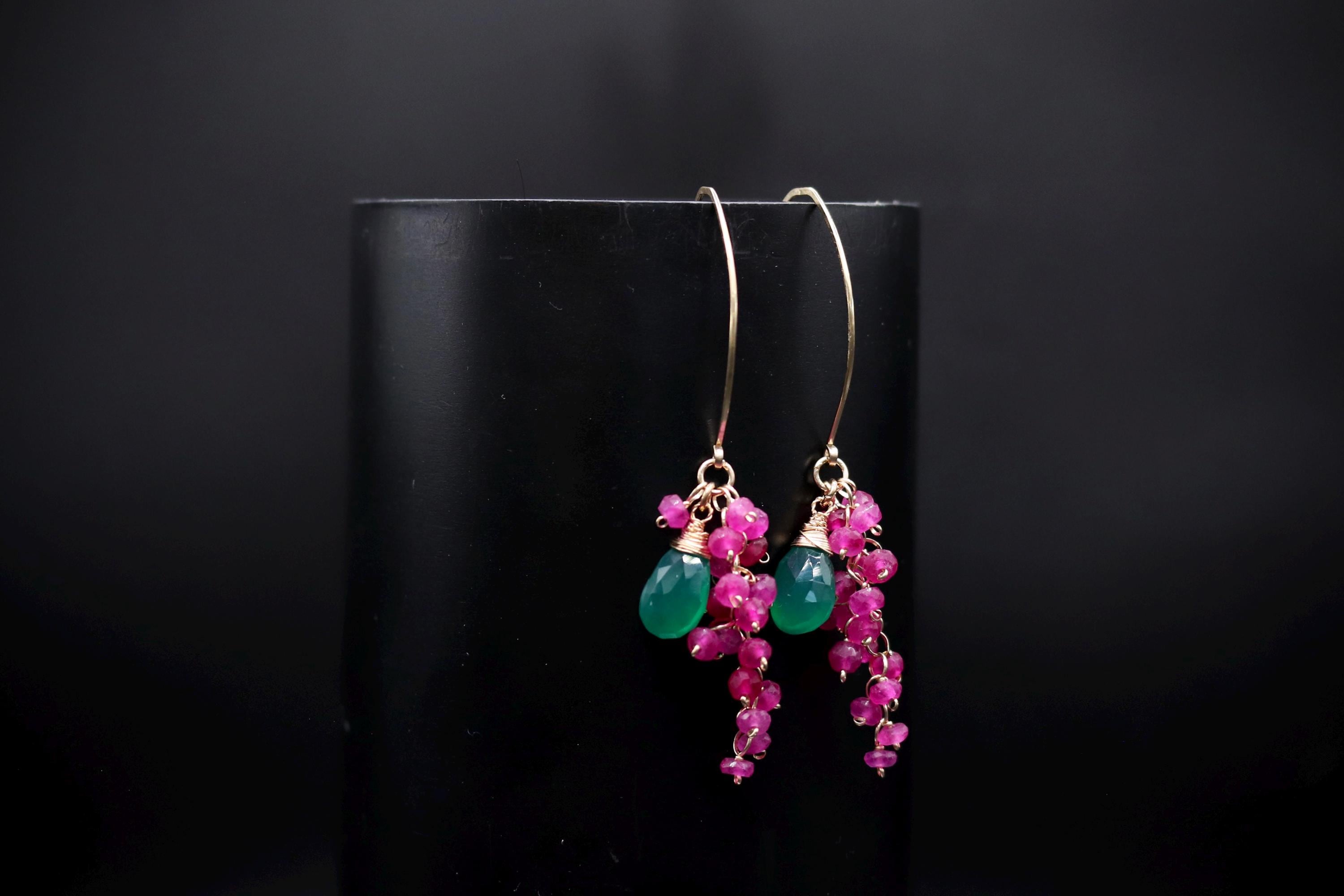 A pair of earrings with green and pink gemstones and dangling charms, displayed against a black background.