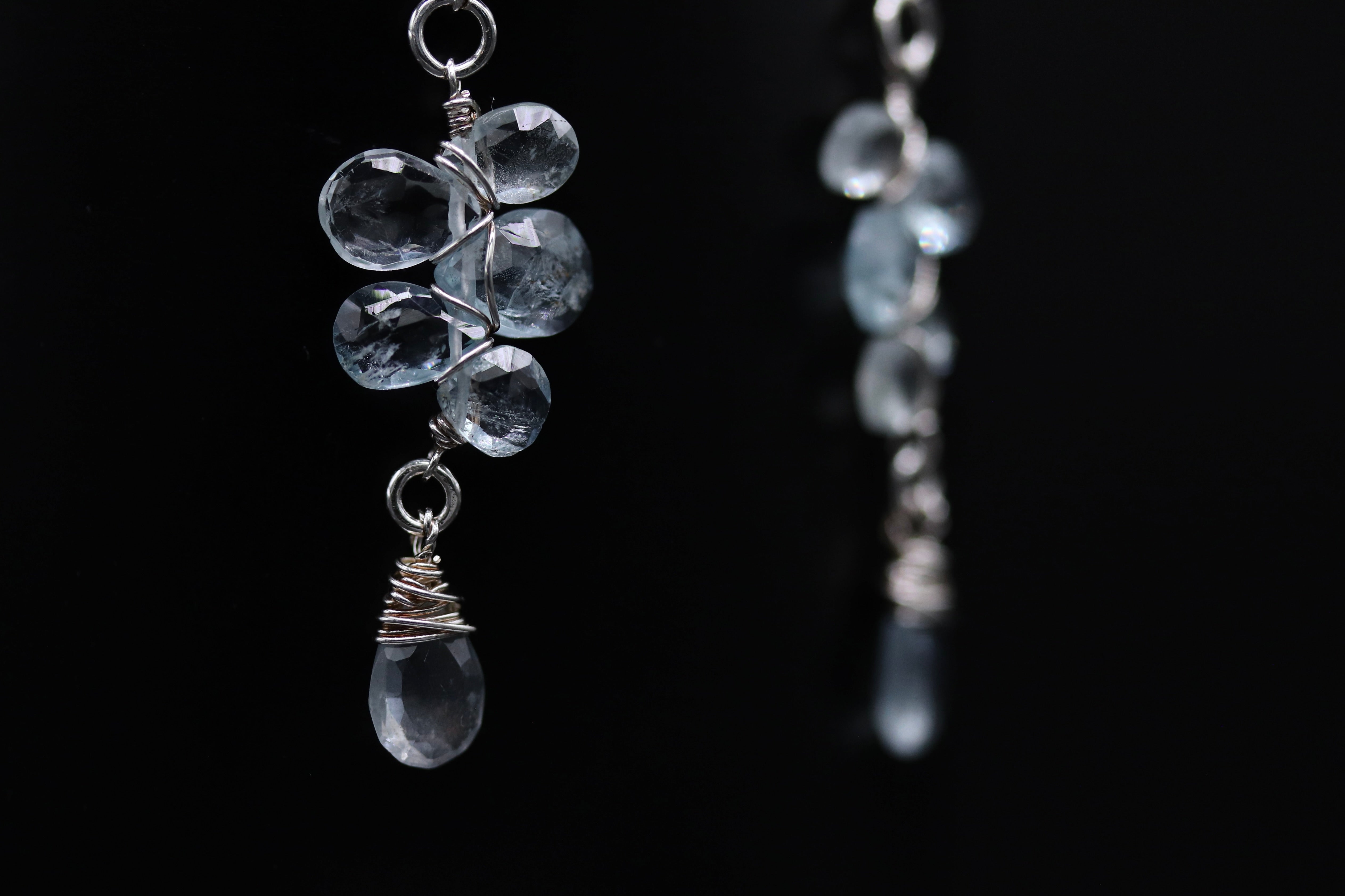 A close-up view of a pair of earrings with clear crystal pendants, suspended from a metal chain against a black background.