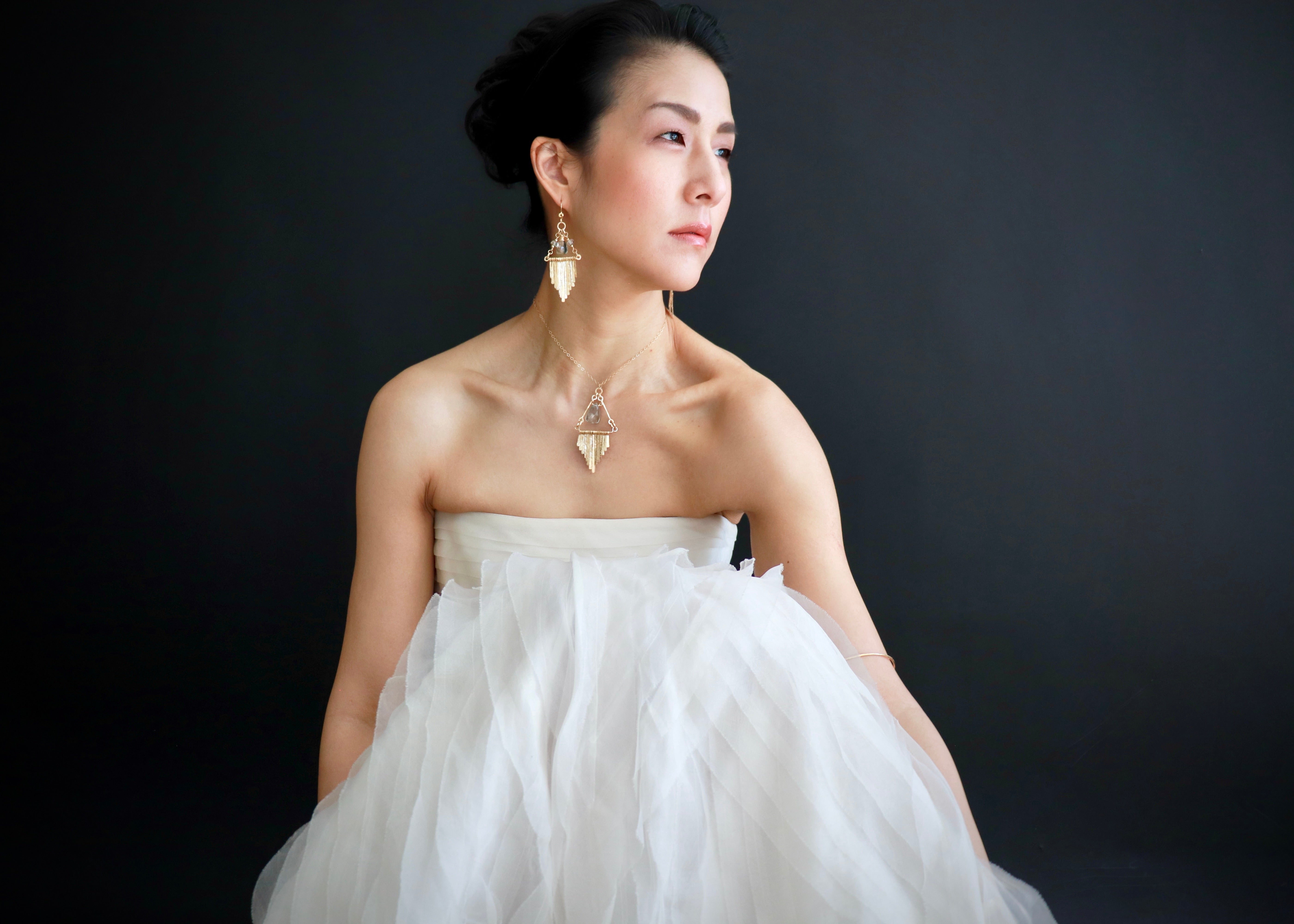 A woman in a white strapless dress with a ruffled skirt stands against a dark background, wearing large gold earrings.