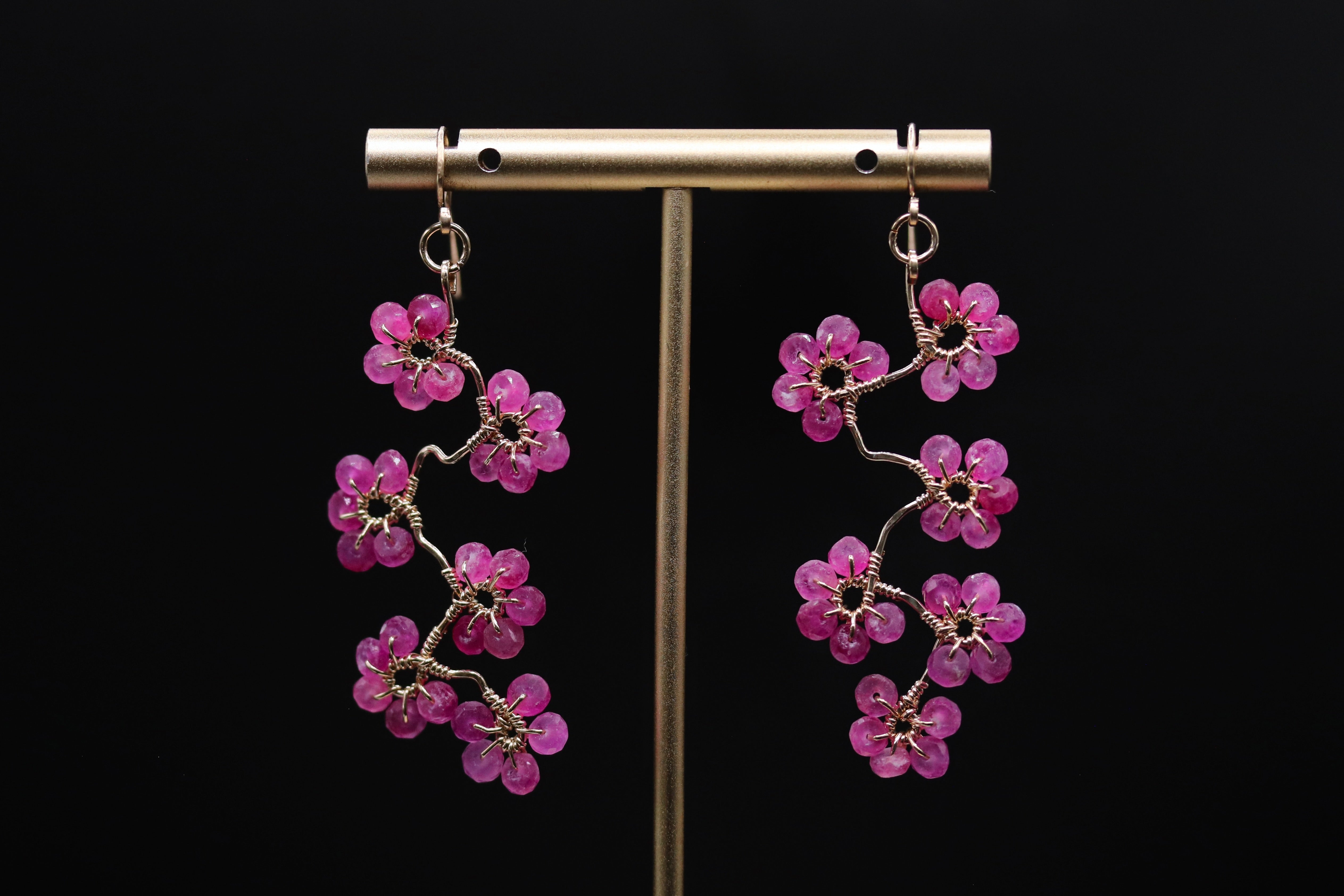 A pair of earrings with pink flower-shaped charms hanging from a metal stand against a black background.