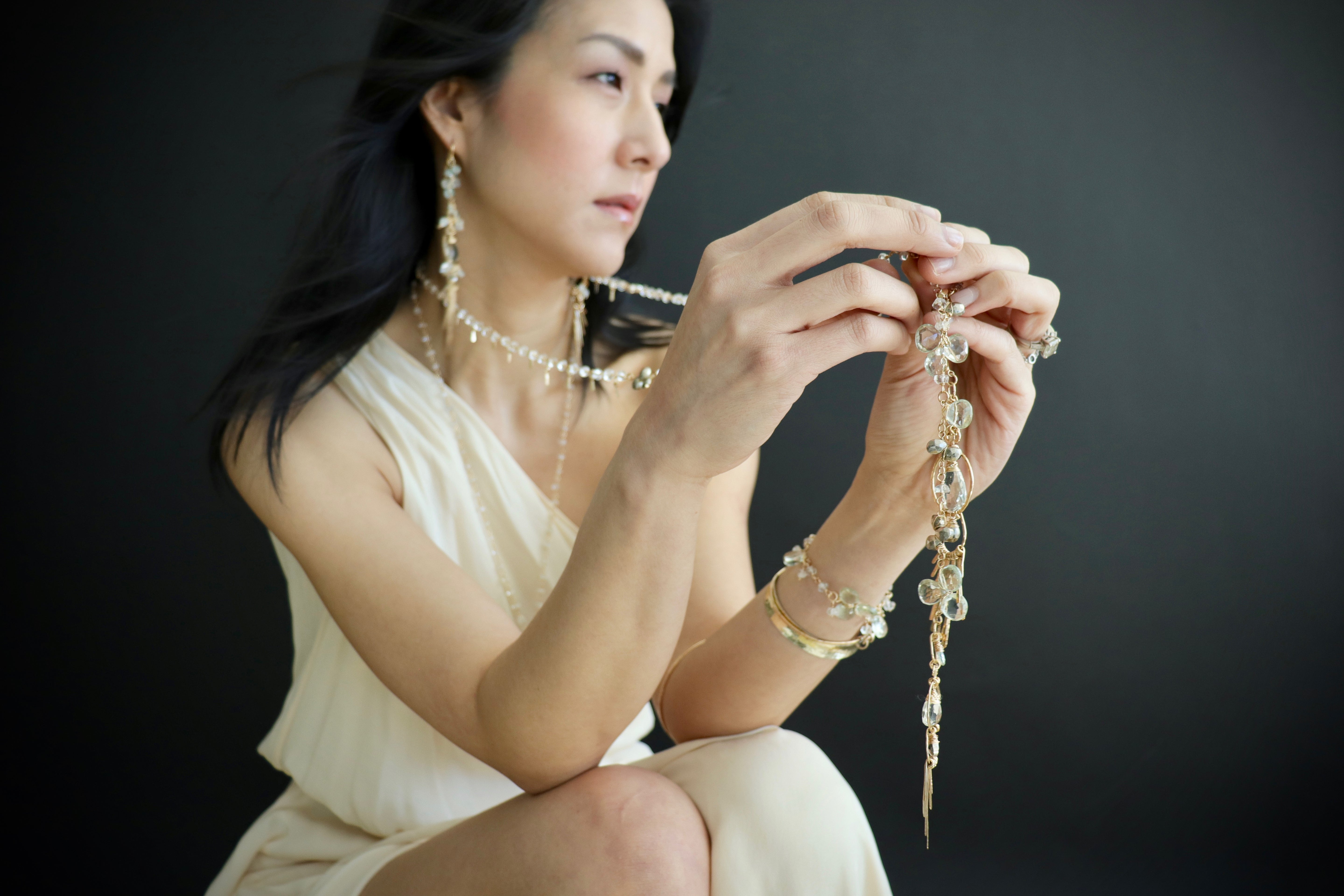 A woman in a white dress is holding a necklace and examining it closely against a dark background.