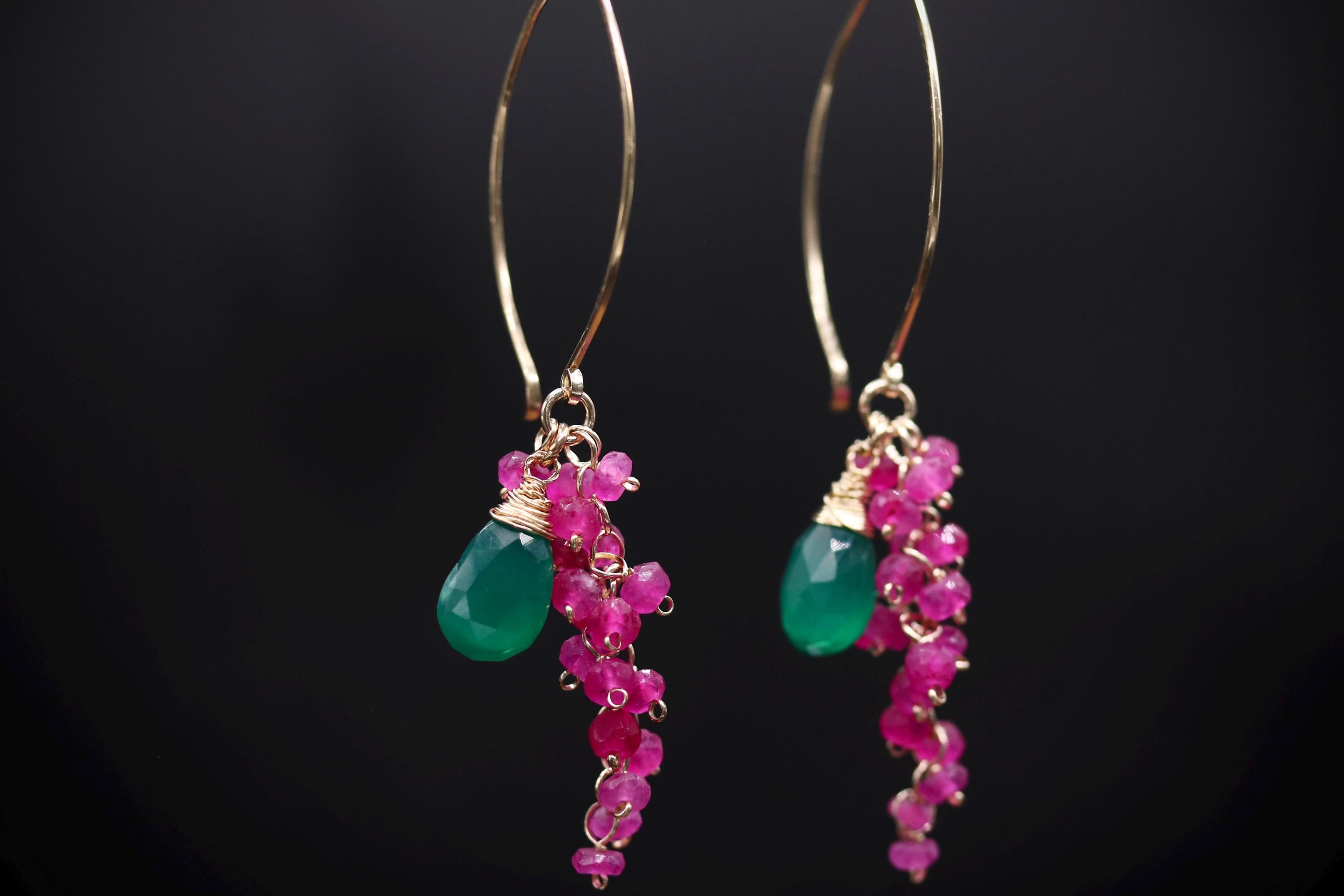 A pair of earrings with a green gemstone and pink gemstone dangling from a gold hook against a black background.
