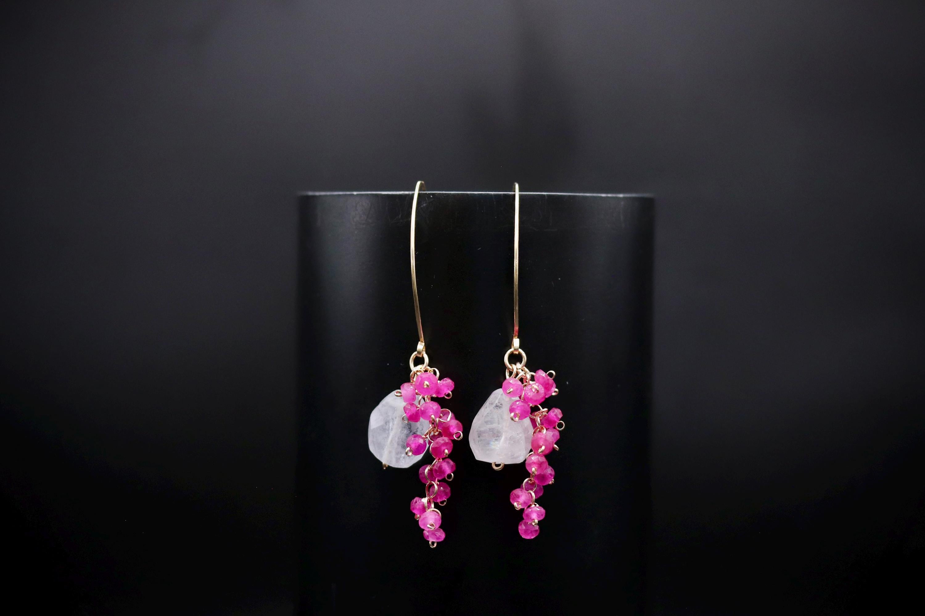 A pair of elegant earrings with a combination of pink and clear gemstones, hanging from a gold hook against a black background.