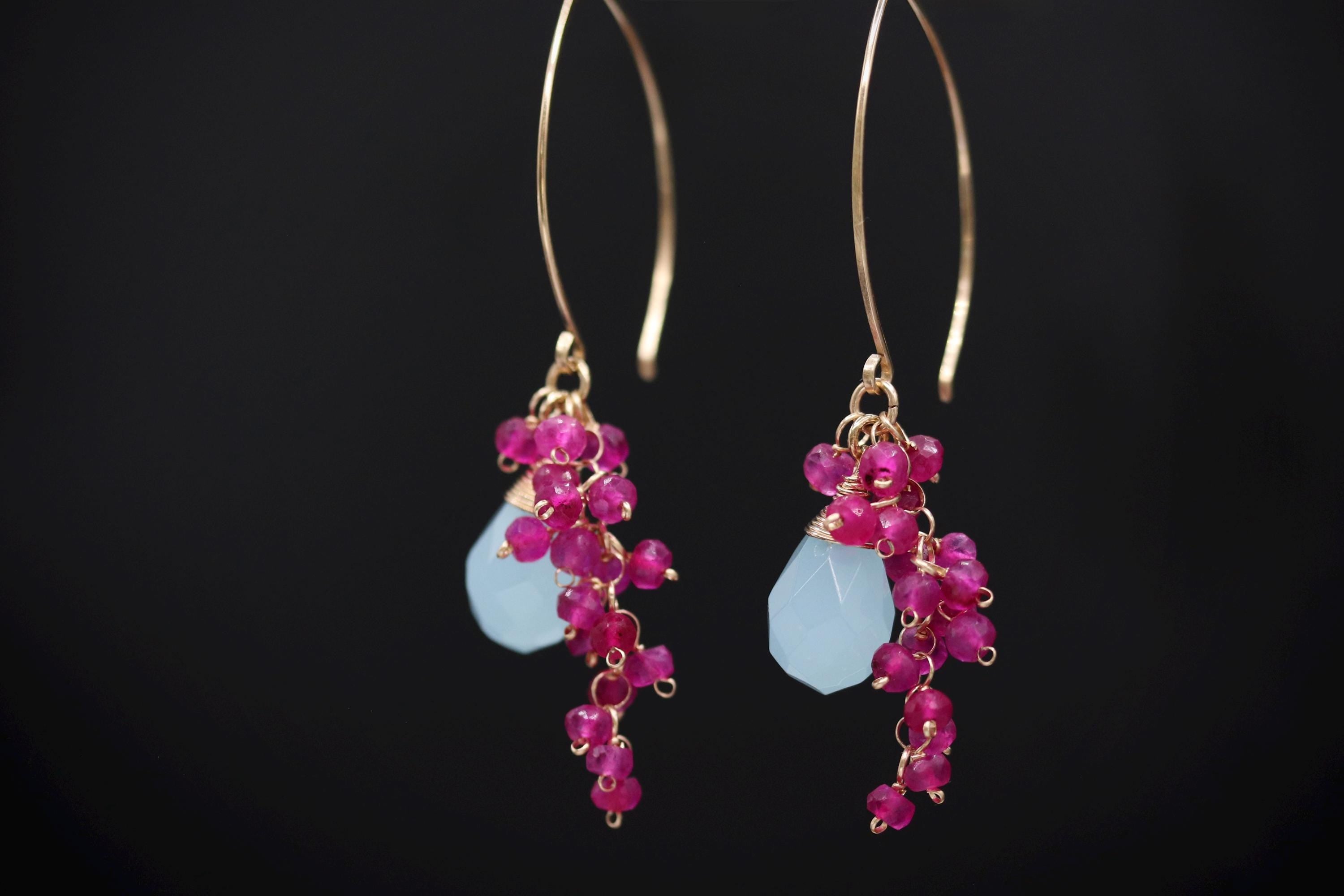 A pair of earrings with a combination of blue and pink gemstones, as well as small pink beads, hanging from a gold hook against a black background.