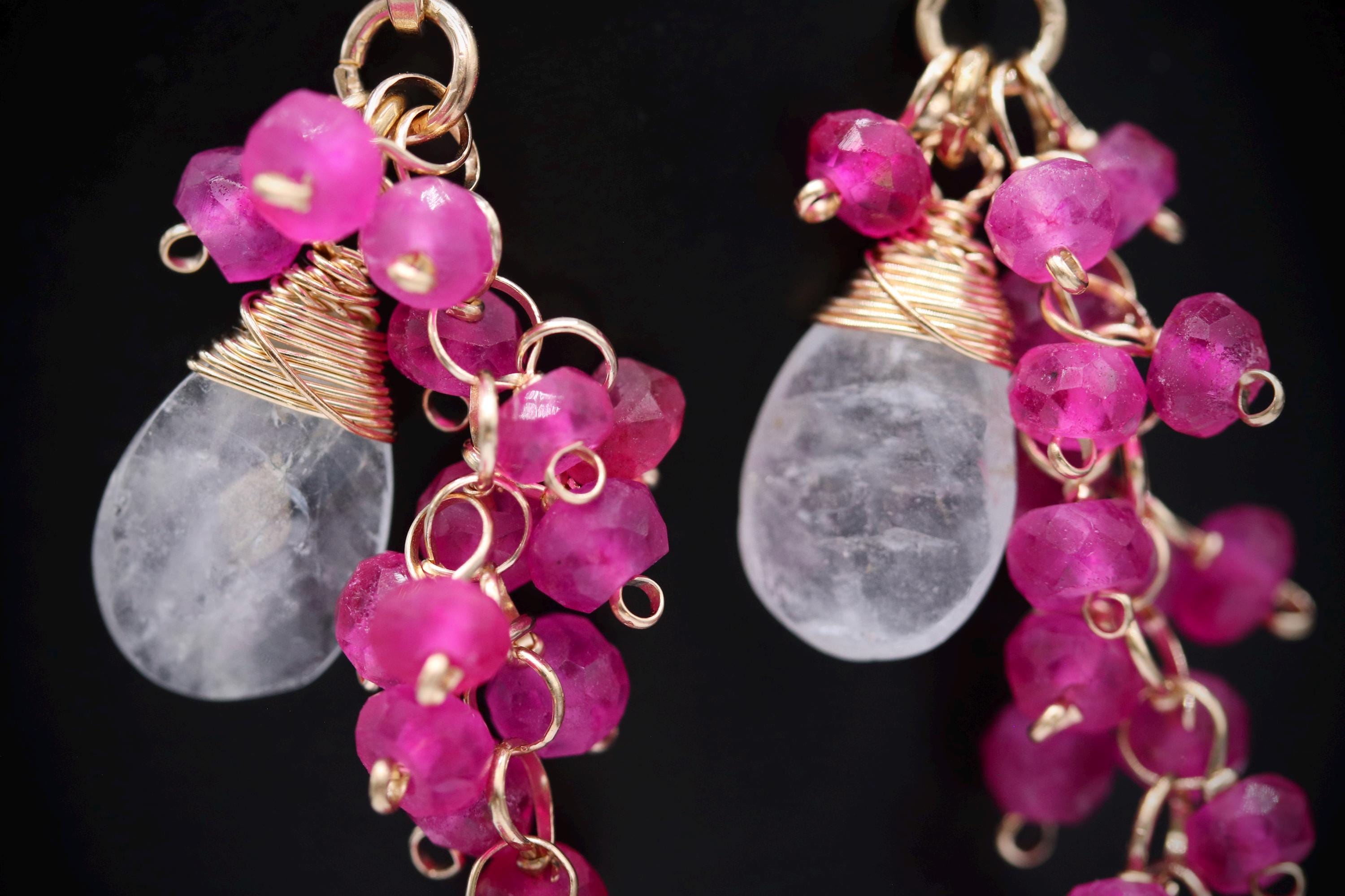 A close-up image of a pair of earrings with clear gemstones and metal chains, featuring a combination of pink and clear gemstones.