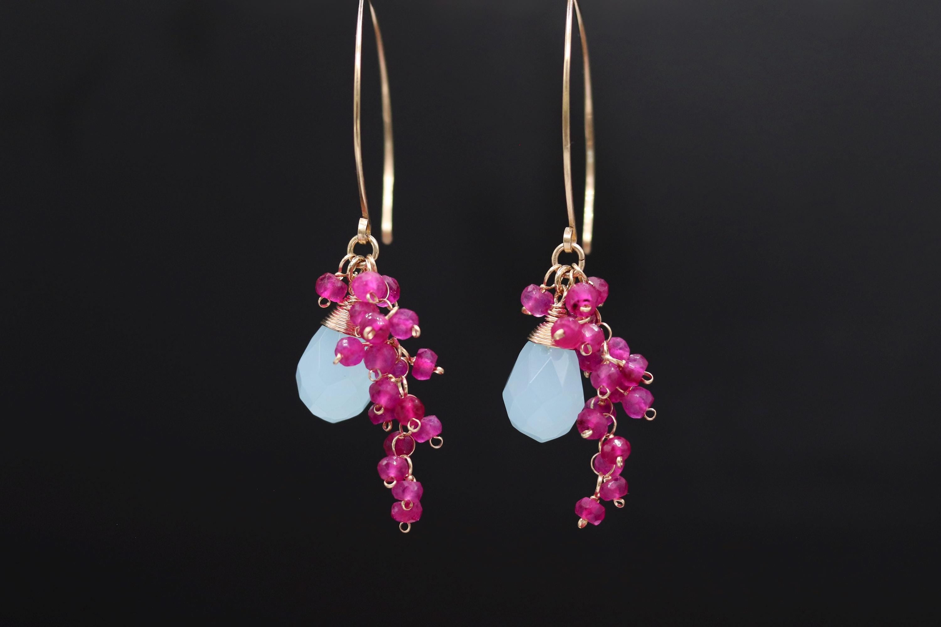 A pair of earrings with a combination of blue and pink gemstones, as well as small pink beads, hanging from a gold hook against a black background.