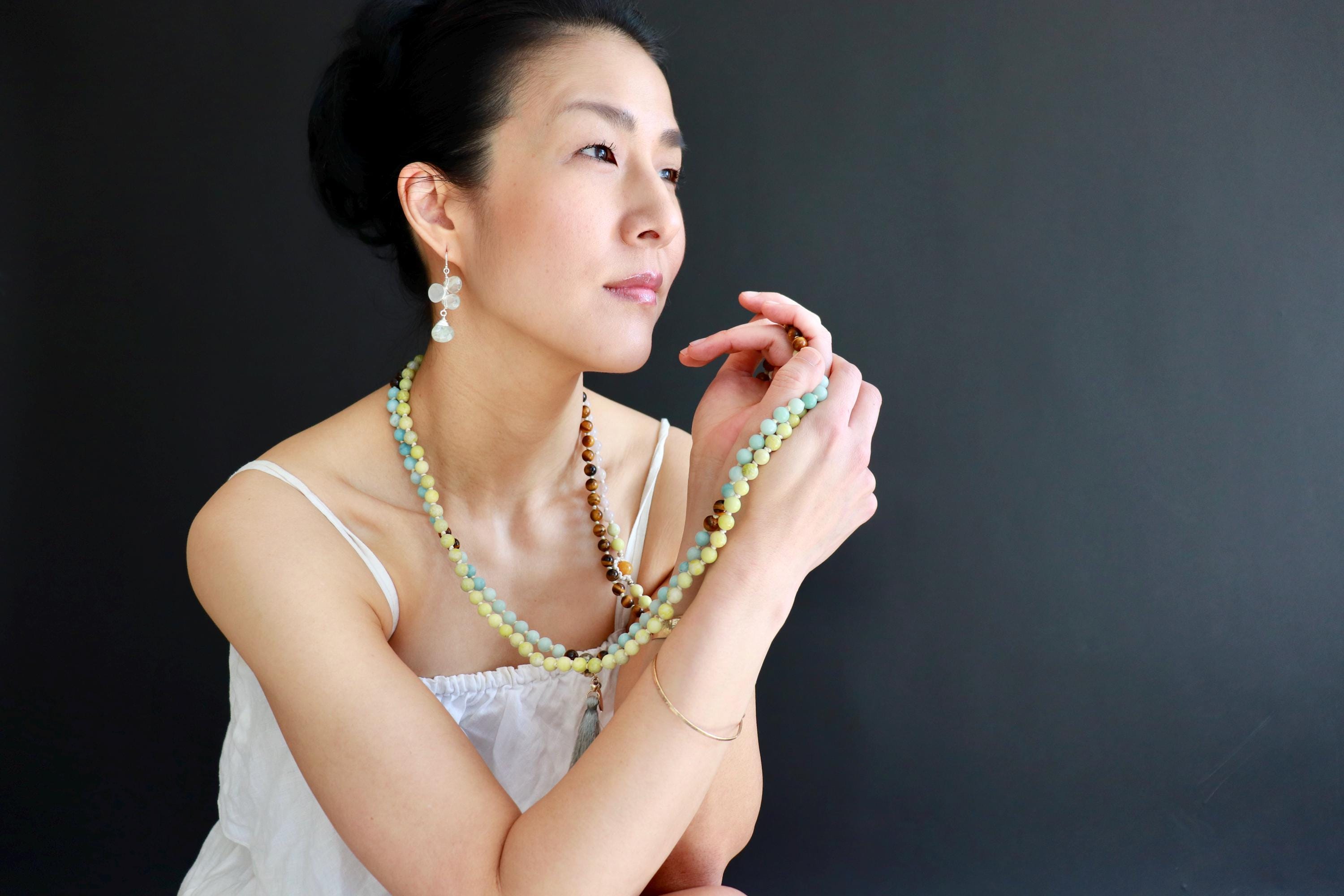 A woman in a white tank top and holding a beaded necklace stands against a dark background, gazing off to the side.
