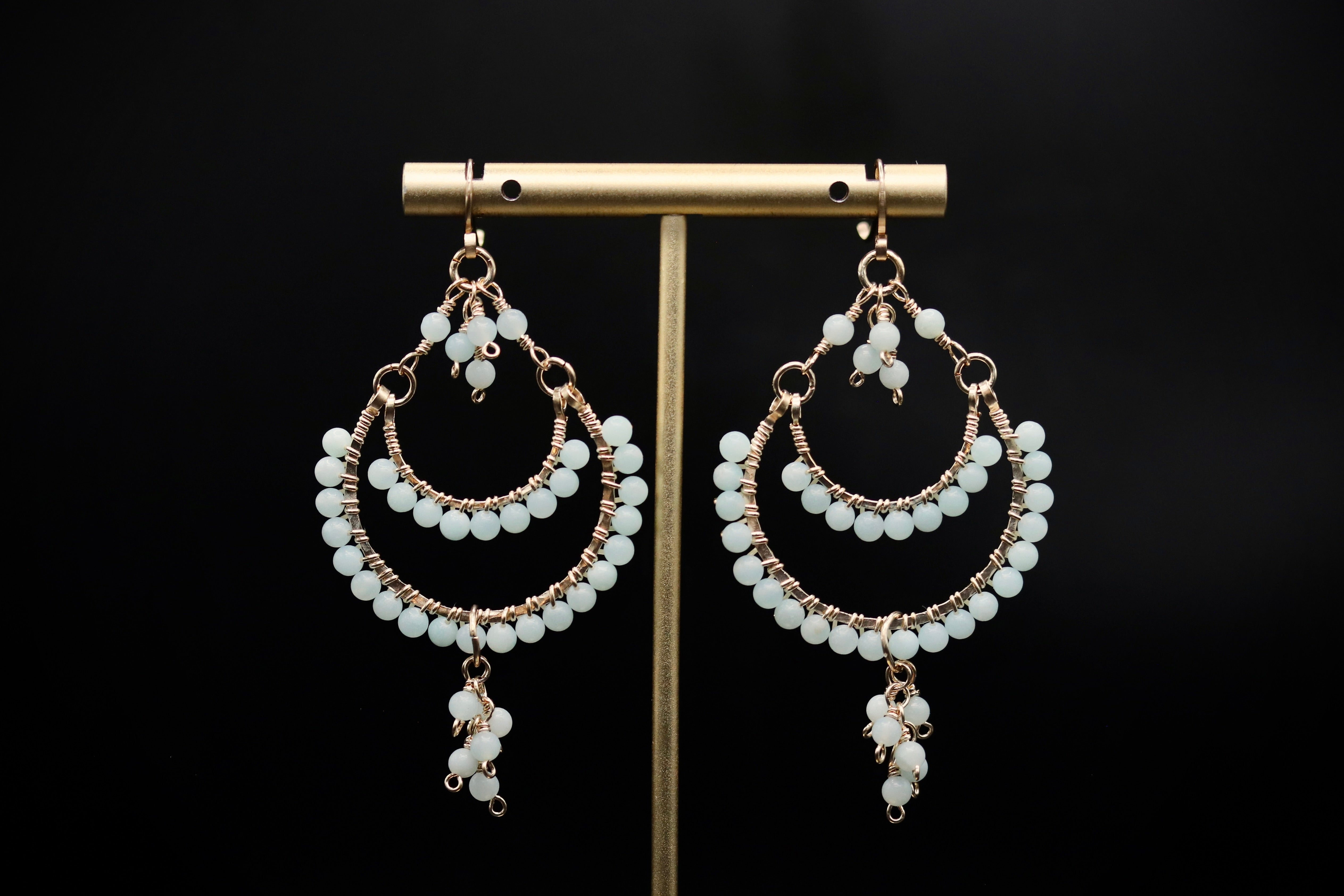 A pair of elegant earrings with white beads and dangling tassels, displayed on a gold stand against a black background.