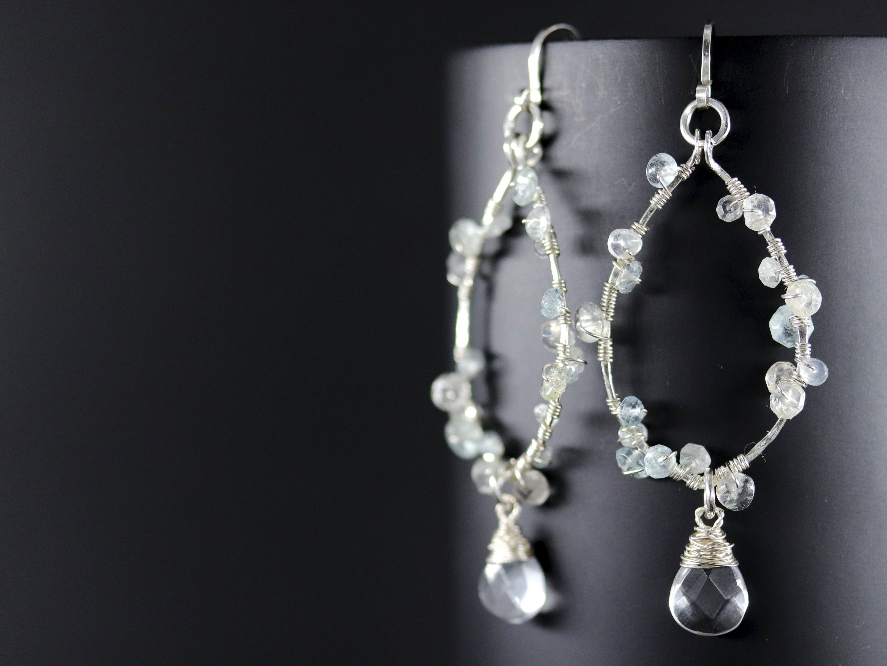 May include: Silver hoop earrings with a teardrop-shaped aquamarine gemstone dangling from each hoop. The hoops are adorned with small aquamarine gemstones.