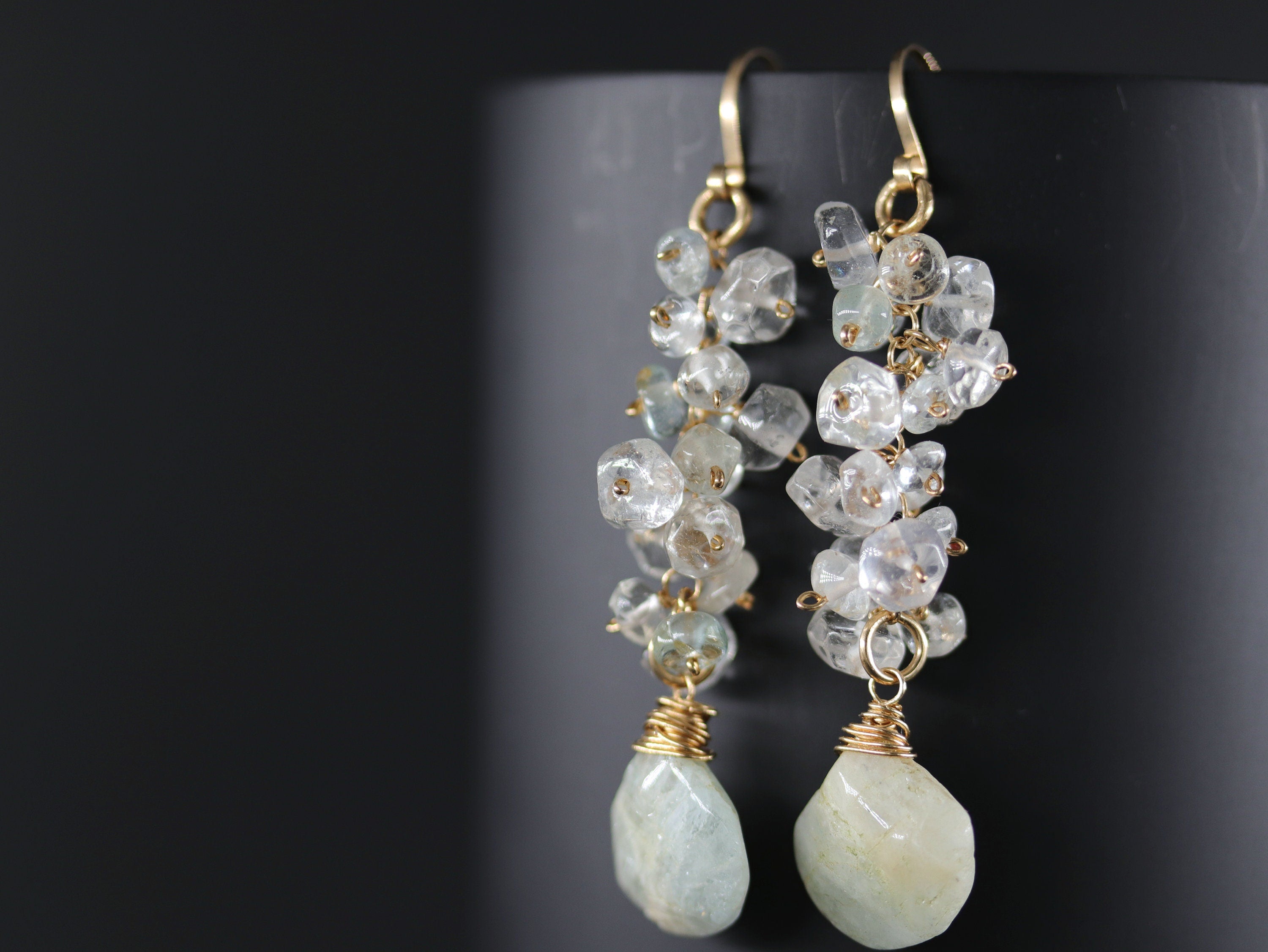 May include: A pair of long, gold dangle earrings with multiple clear and light blue gemstone beads. The earrings have a teardrop-shaped gemstone at the bottom.