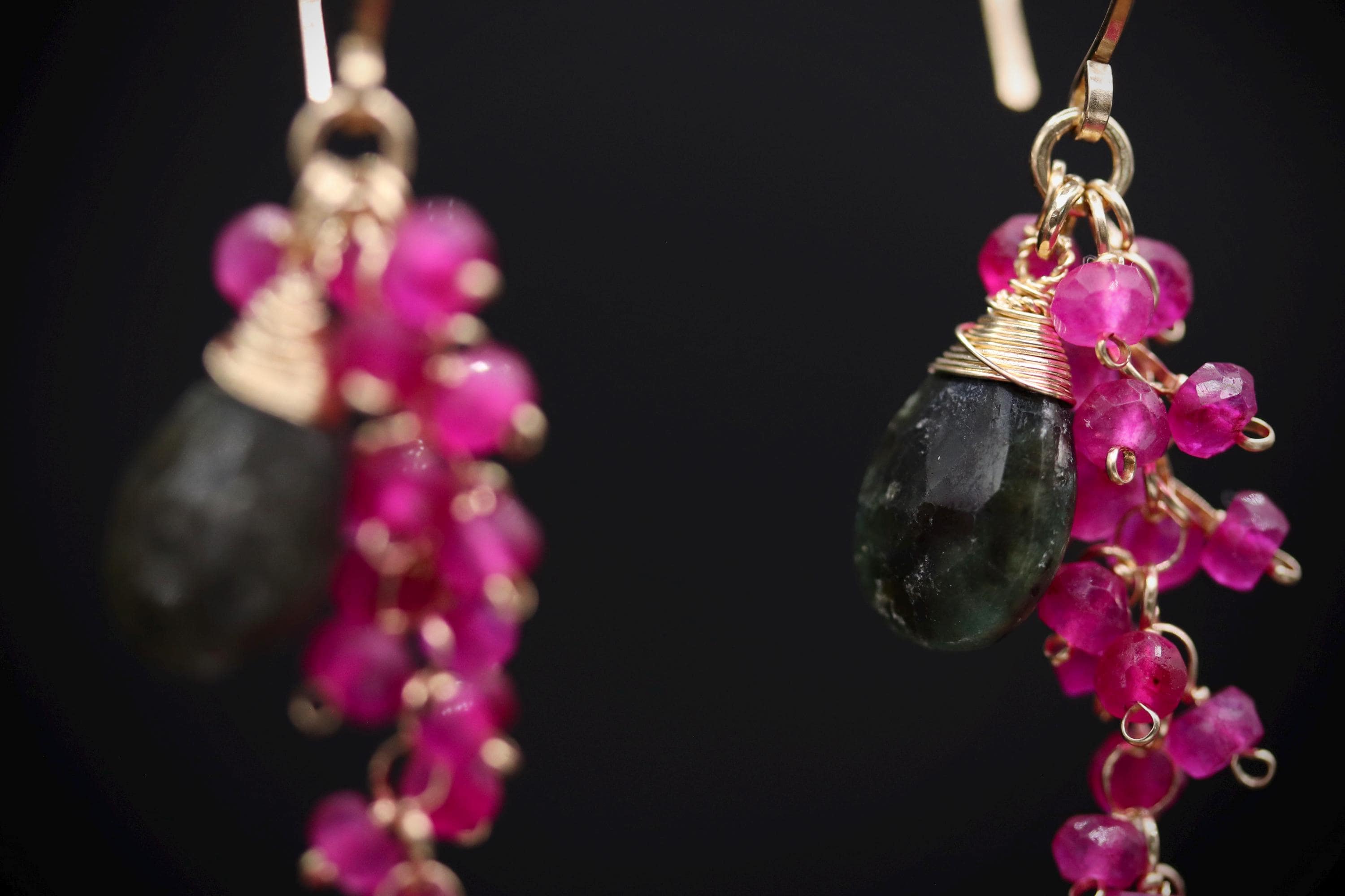 A close-up view of a pair of earrings with a combination of green and pink gemstones and dangling charms.