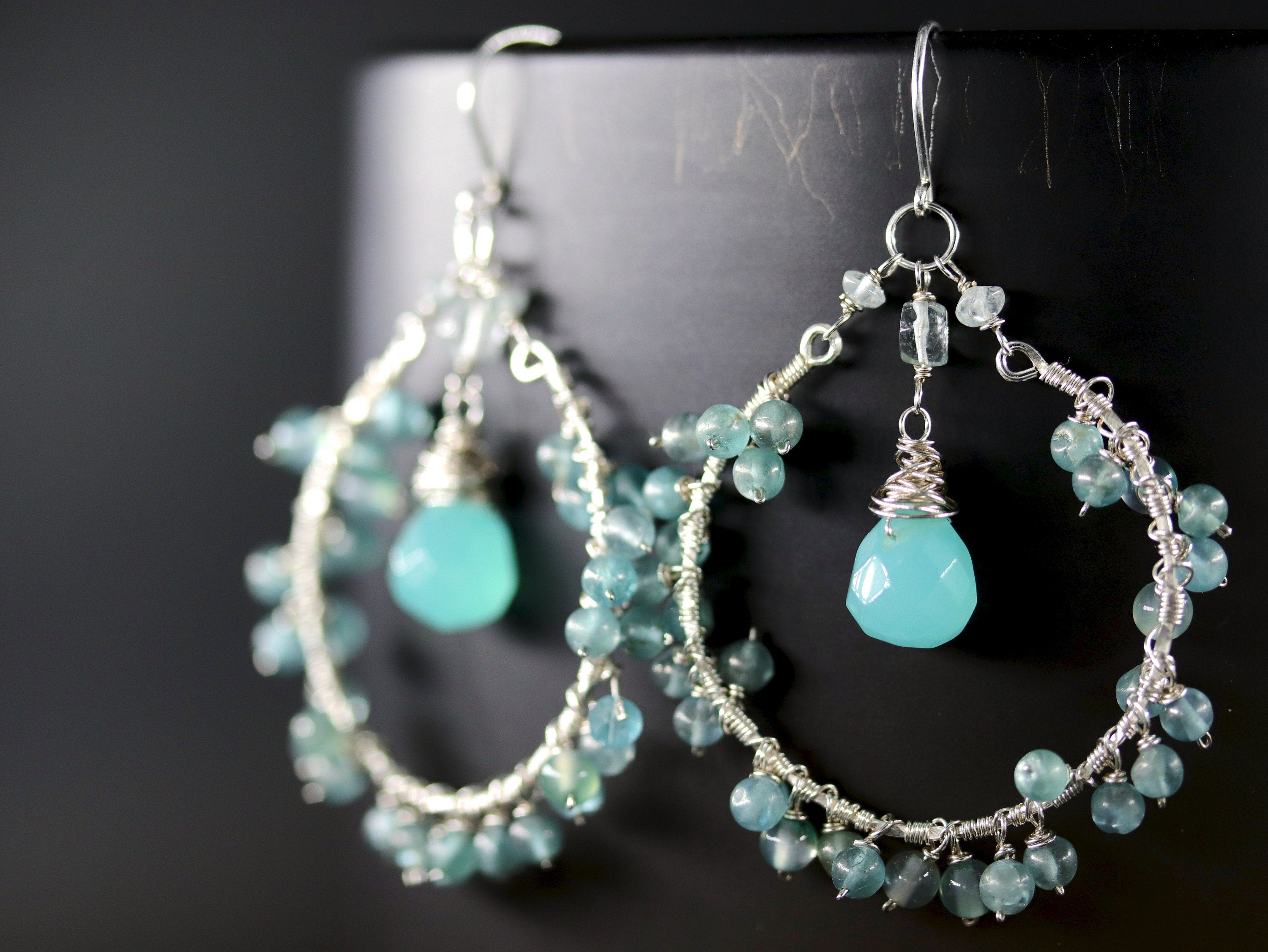 May include: Silver hoop earrings with a large light blue gemstone and smaller blue gemstones around the hoop.