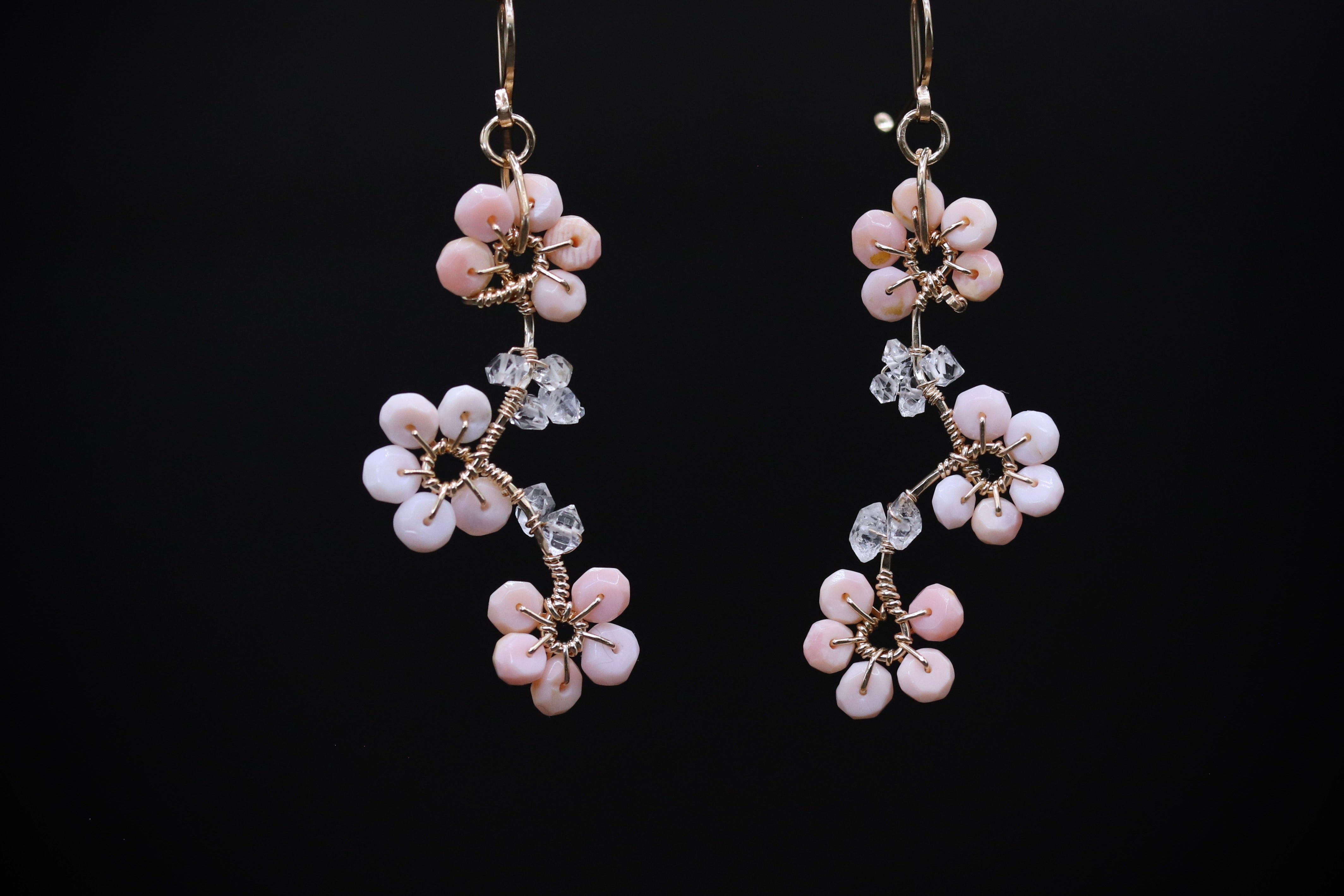 A pair of delicate earrings with pink flowers and clear gemstones against a black background.