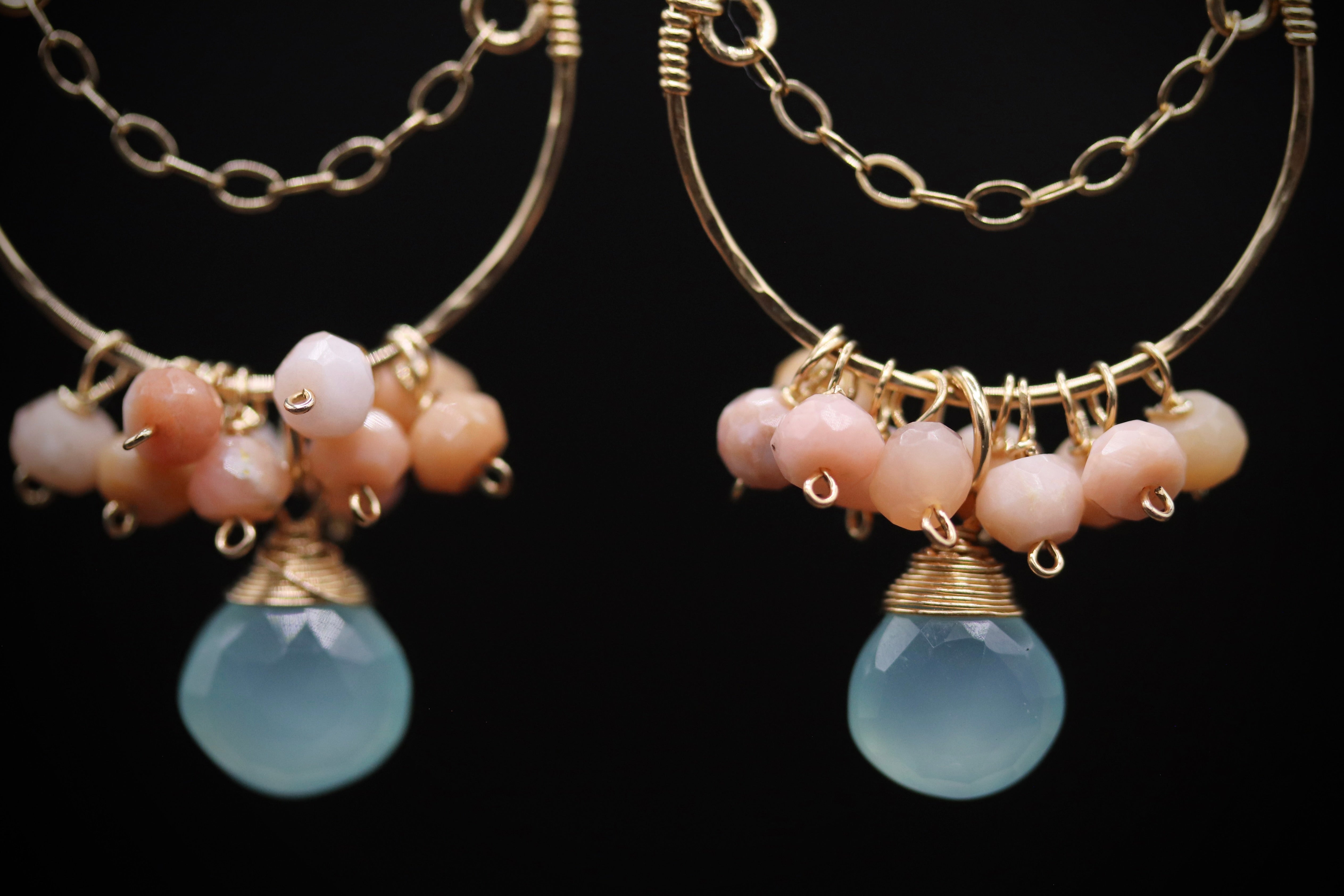 A pair of gold hoop earrings with a blue gemstone and small beads hanging from the chain.