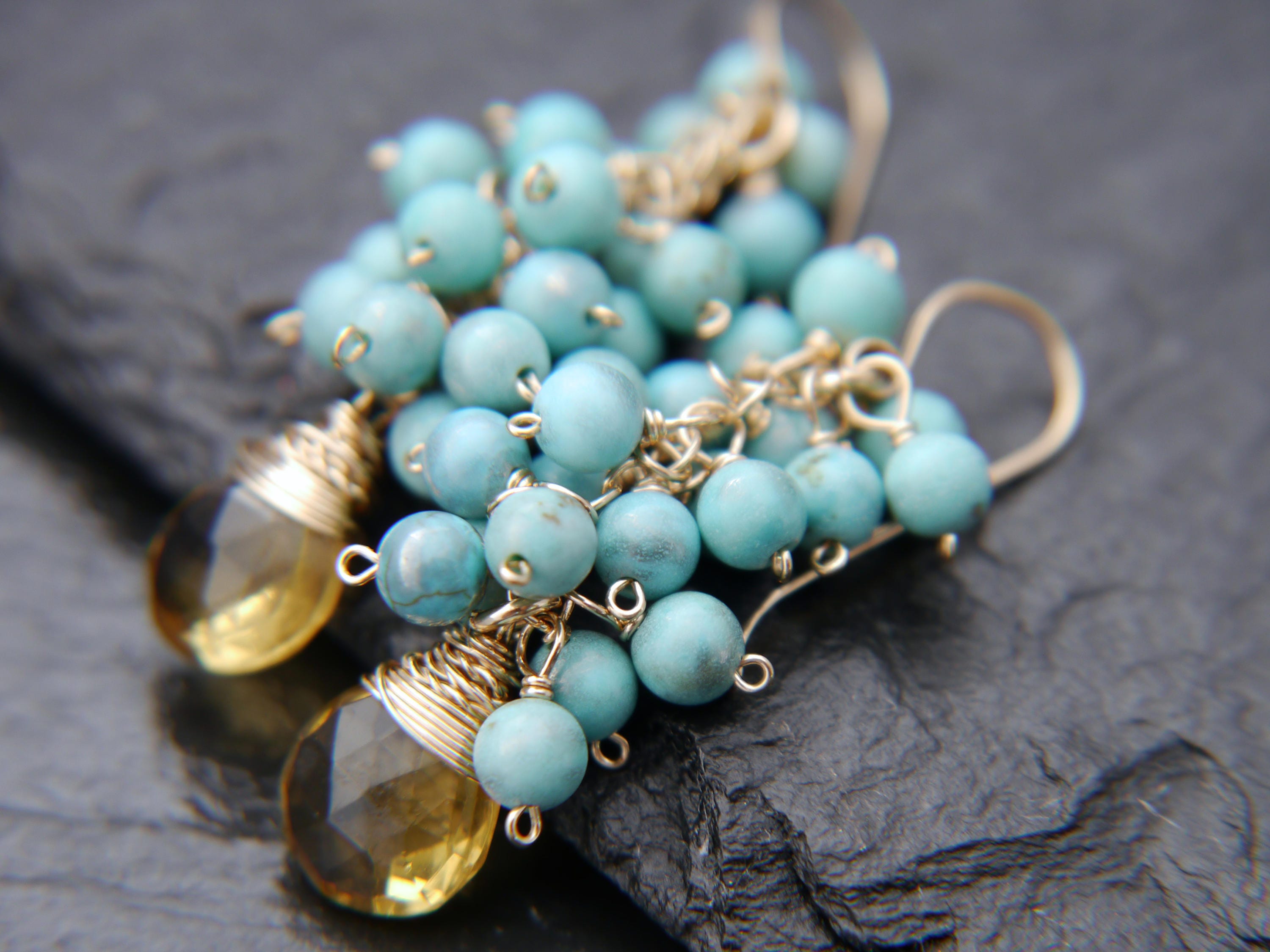 May include: Close-up of dangle earrings featuring clusters of small, round, turquoise beads. The earrings also have faceted, teardrop-shaped, yellow-gold gemstones. The metal components are gold-toned.