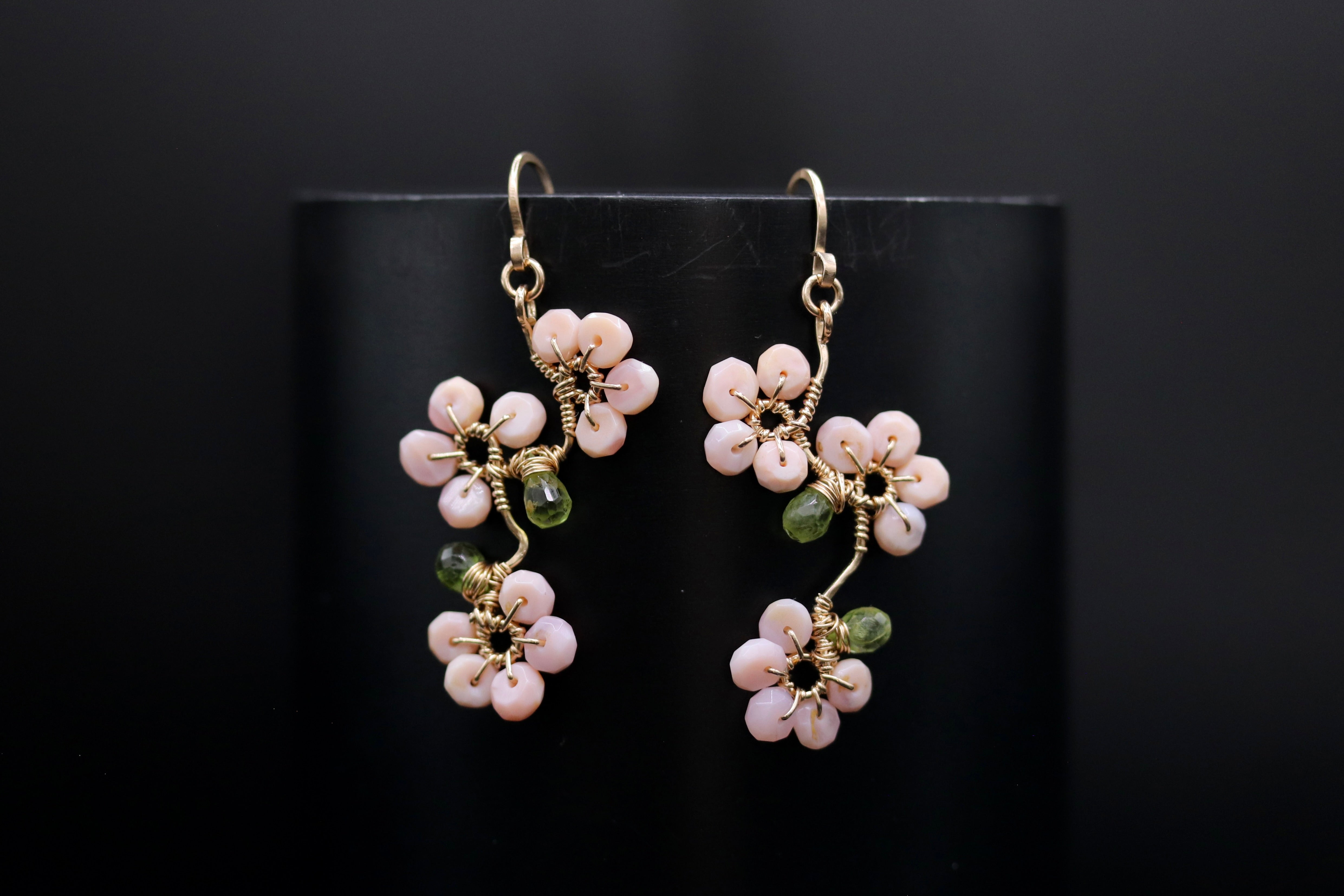 A pair of delicate earrings with pink and green flower-shaped charms, hanging from gold hooks against a black background.