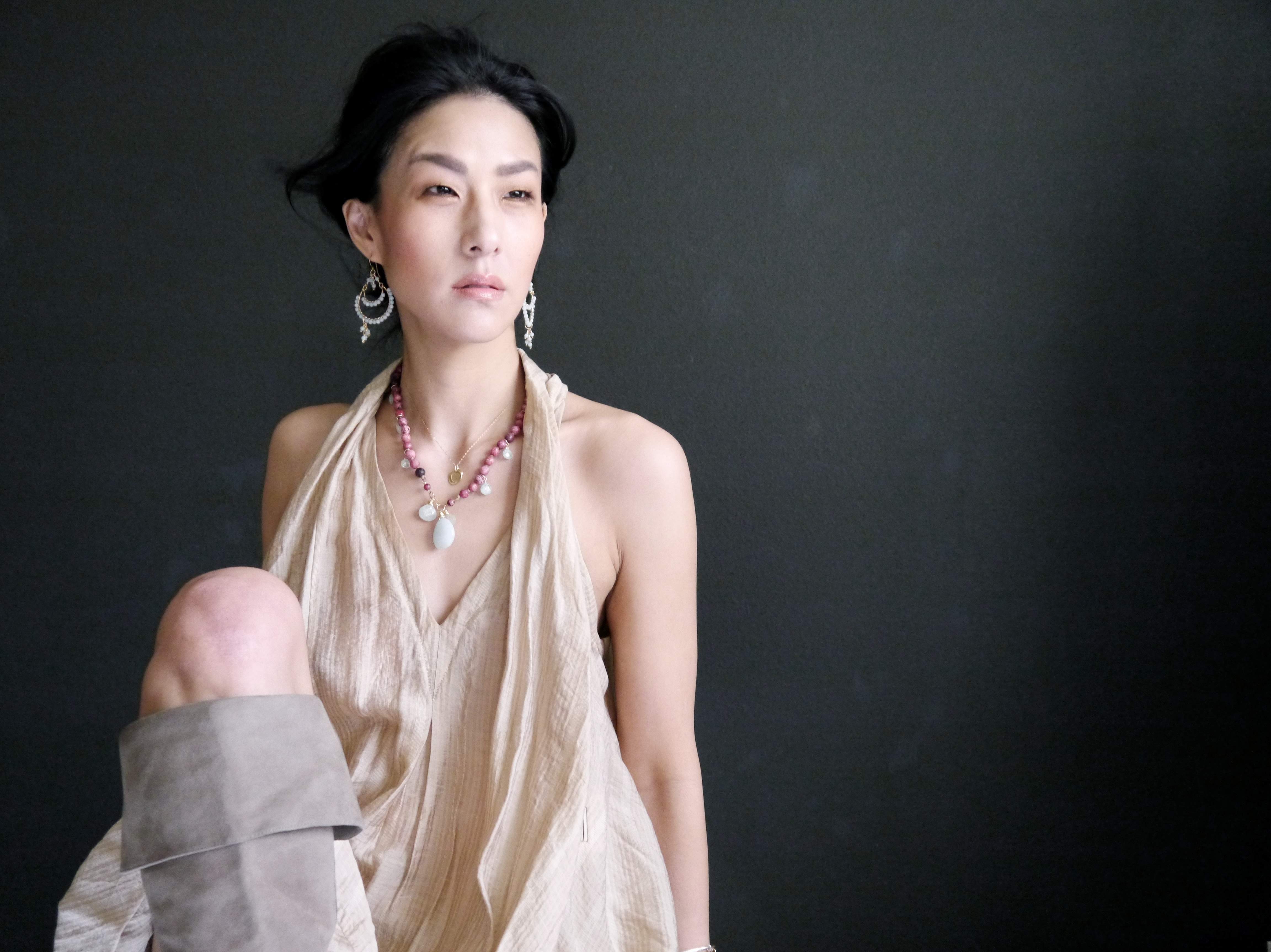 A woman wearing a beige dress and earrings stands against a dark background, looking off to the side with a serious expression.