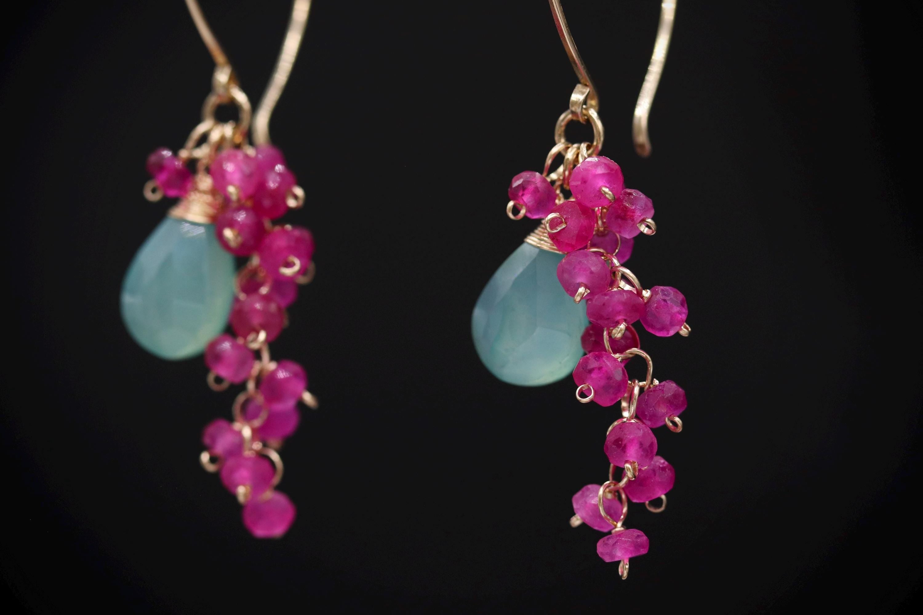 A pair of earrings with teal-colored stones and pink gemstone beads hanging from the ear hooks against a black background.
