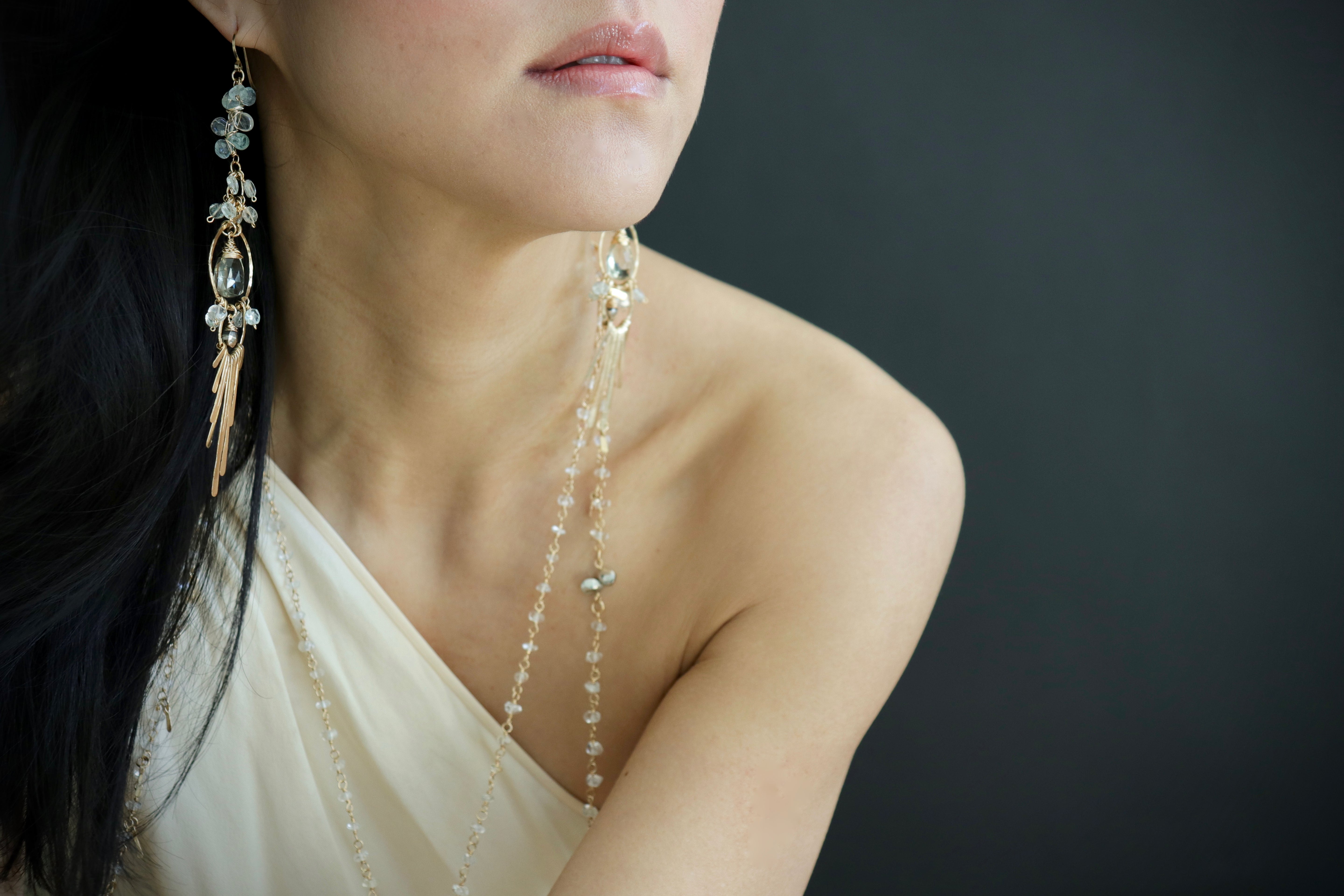 A close-up of a woman's face wearing large, dangling earrings and a necklace against a dark background.