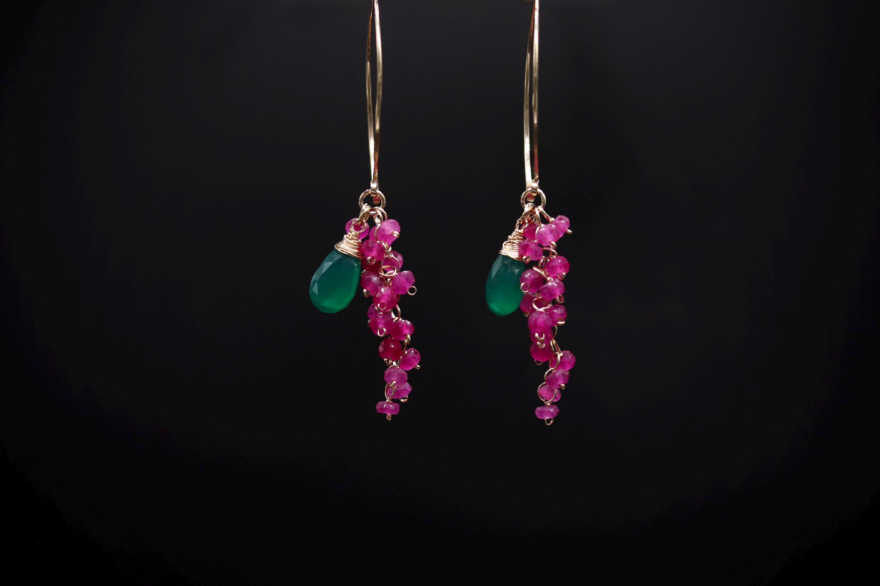 A pair of earrings with green and pink gemstones hanging from a gold hook against a black background.