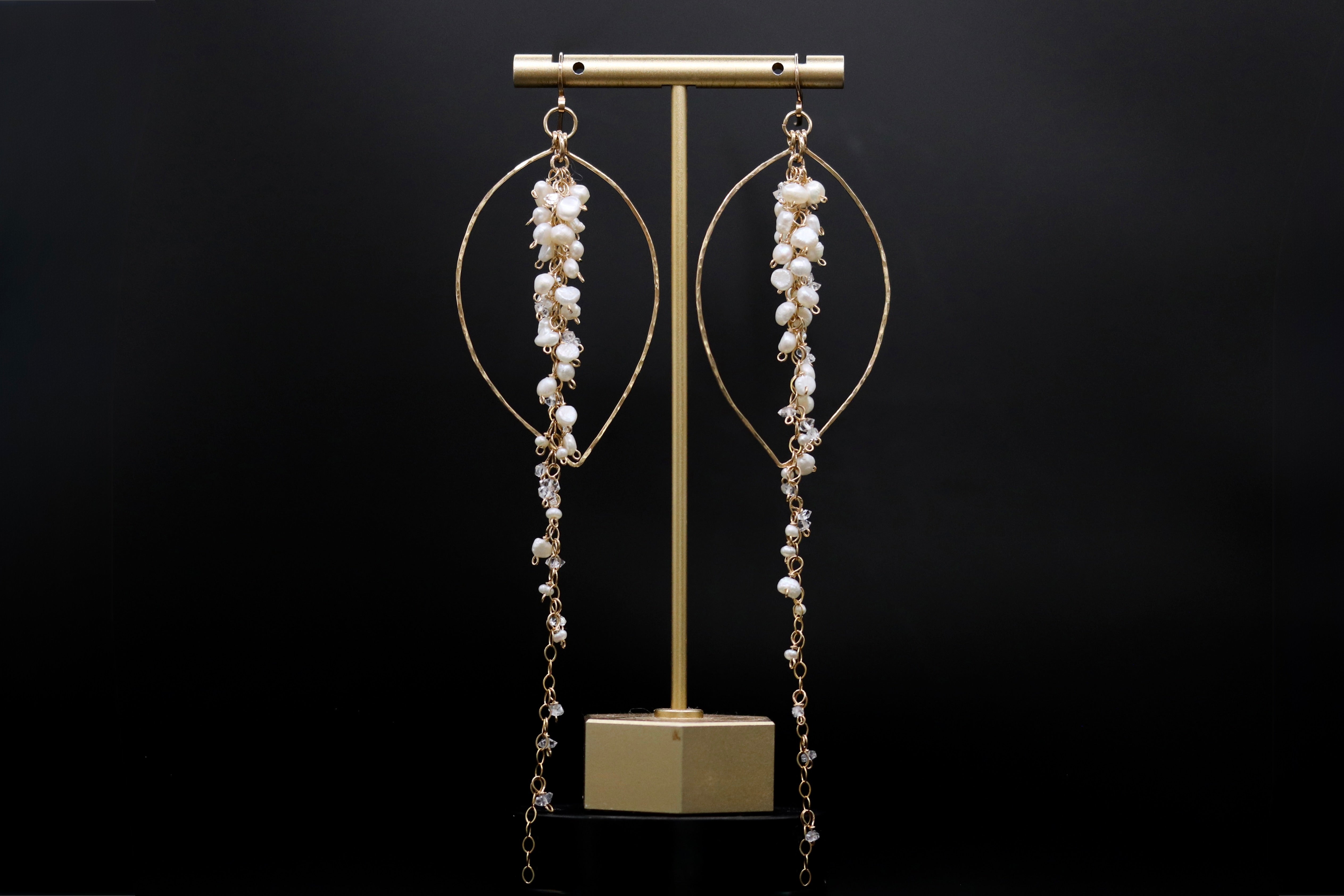 A pair of elegant earrings with long, dangling chains and small white pearls or crystals hanging from them, displayed on a gold stand against a black background.